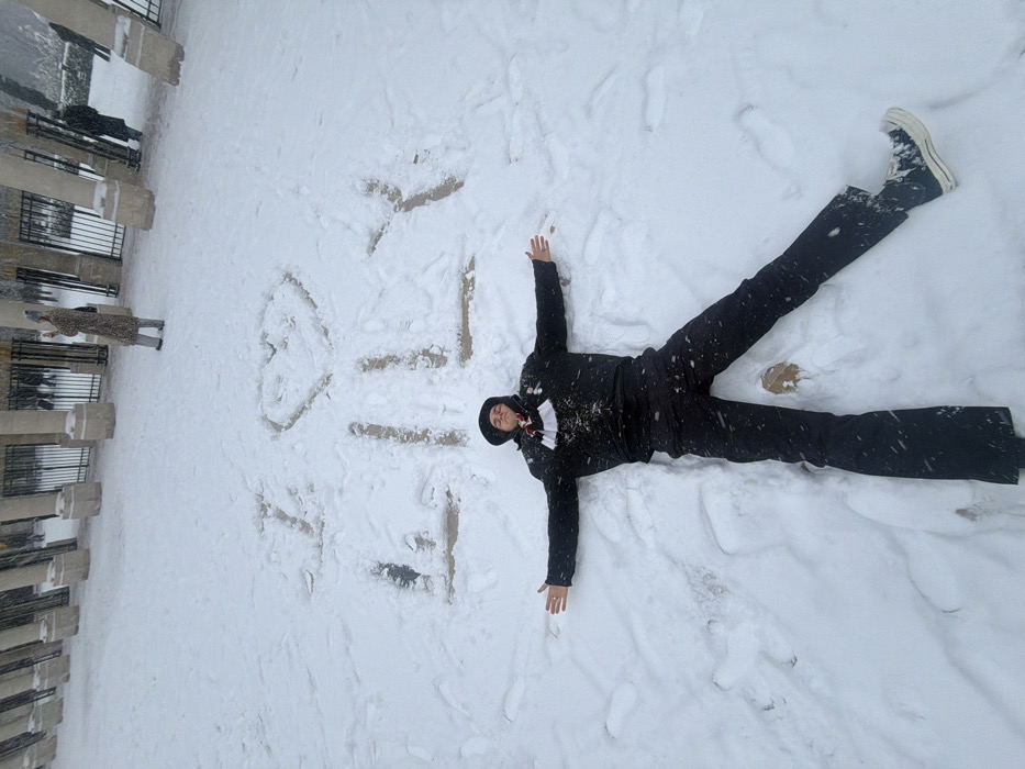 The image captures a serene and playful winter scene in an outdoor setting in Paris, France. In the foreground, a person is lying on their back in a thick blanket of fresh, white snow, with arms and legs outstretched, actively engaged in making a snow angel. They are dressed warmly in dark winter attire, including a black jacket, pants, and a dark hood or beanie. The person's face is partially visible, showing a calm or slightly smiling expression, suggesting enjoyment of the moment. Snow specks are visible on their clothing, indicating either ongoing snowfall or recent movement in the snow.
The snow around the person clearly shows the distinctive imprints of a snow angel's "wings" and body. To the left, in the mid-ground, another person is partially visible, standing upright and looking away from the main subject, towards a long, dark metal fence or railing that runs horizontally across the left side of the frame. This fence borders a paved area or pathway, beyond which faint architectural elements and bare trees are discernible, consistent with a public park or garden in an urban environment like Paris. The ground is entirely covered in undisturbed snow, suggesting a recent snowfall. The lighting is soft and diffused, characteristic of an overcast daytime. There is no visible text in the image.