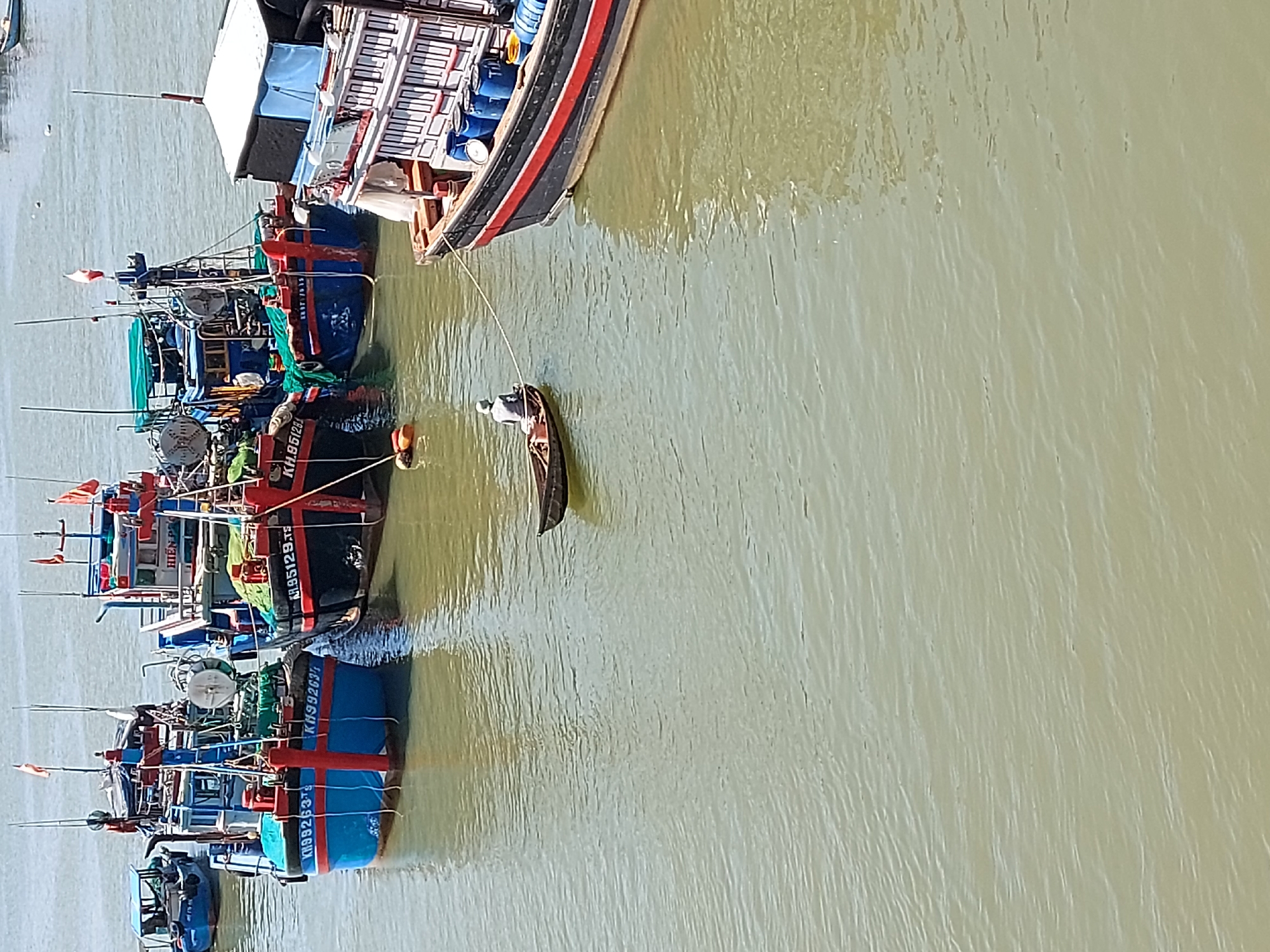 The image depicts a body of water, likely a harbor or bay, with several boats. In the foreground, a large fishing vessel with a red and black hull is docked. It has several blue barrels on its deck. To the left of this vessel, a small, dark-colored rowboat is in motion, carrying a single individual wearing a light-colored shirt and hat. This rowboat is tethered to the larger fishing vessel by a rope. Behind these boats, a fleet of larger fishing boats, predominantly blue and red, are moored. These boats have various structures on their decks, including nets and equipment. The water is a murky, greenish-brown color, with ripples indicating gentle movement. The sky is bright and appears to be a sunny day. The location is identified as the city of Bac Nha Trang Ward, Vietnam.