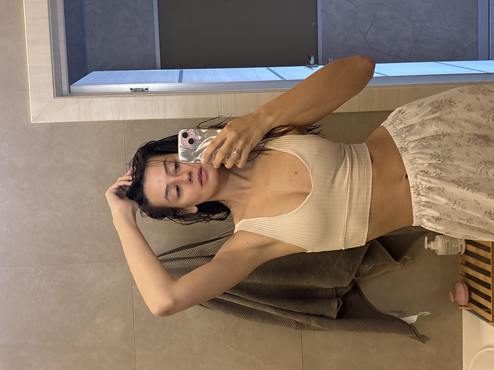The image is a mirror selfie of a young woman in a modern bathroom setting, rotated 90 degrees counter-clockwise. The woman, appearing in her 20s or 30s, has wet, dark hair and is wearing a light-colored ribbed crop top and loose white shorts or pajama pants with a subtle grey floral pattern. She holds a silver-toned smartphone with a light pink/clear case up to her face, capturing her reflection. A ring is visible on her left ring finger. Her right hand is raised, gently touching her hair.
The background features large light grey/beige wall tiles. In front of her, a dark grey/brown towel is draped over a surface, and a lighter-colored towel is visible below it. To the right, a white pump bottle, a pink circular object (possibly a sponge), and a wooden slatted caddy are placed on a surface. The mirror reflects an area behind her, which appears to be a window or an architectural opening, showing a dark interior beyond with some diffused light, suggesting an indoor evening or night setting. The lighting is soft and artificial. No distinct text is clearly legible on any visible objects, except for blurry text on the pump bottle. The implied location context is Krasnoyarsk, Russia.
