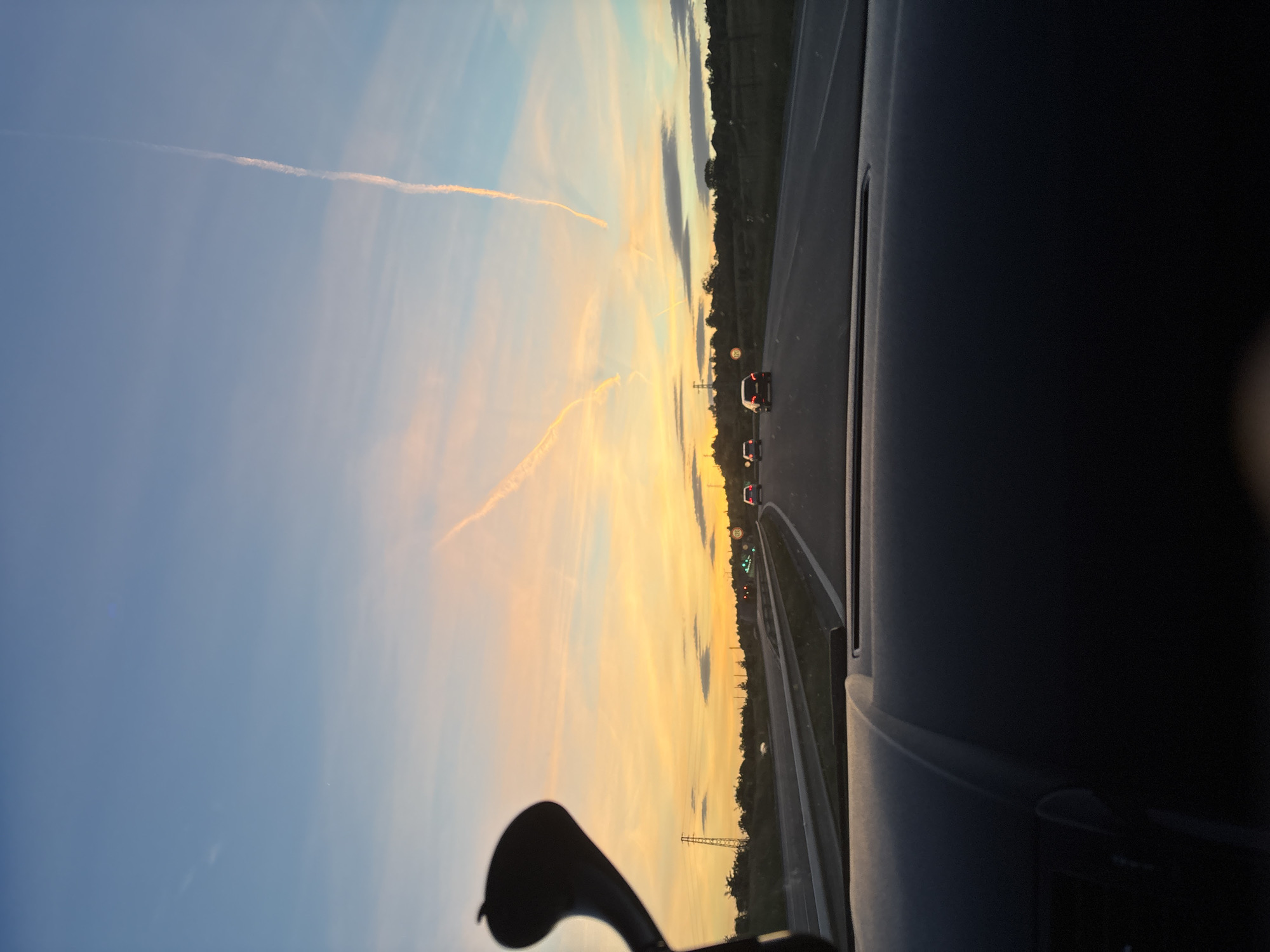 The image depicts a view from inside a moving vehicle, likely a car, traveling on a highway. The sky is visible through the windshield, displaying a sunset with a mix of blue, orange, and yellow hues, and contrails from airplanes. In the distance, there are other cars on the road ahead, with their red taillights visible. On the right side of the image, the dashboard of the car is visible, as well as trees in the distance. The location is Dragichevo, Bulgaria.