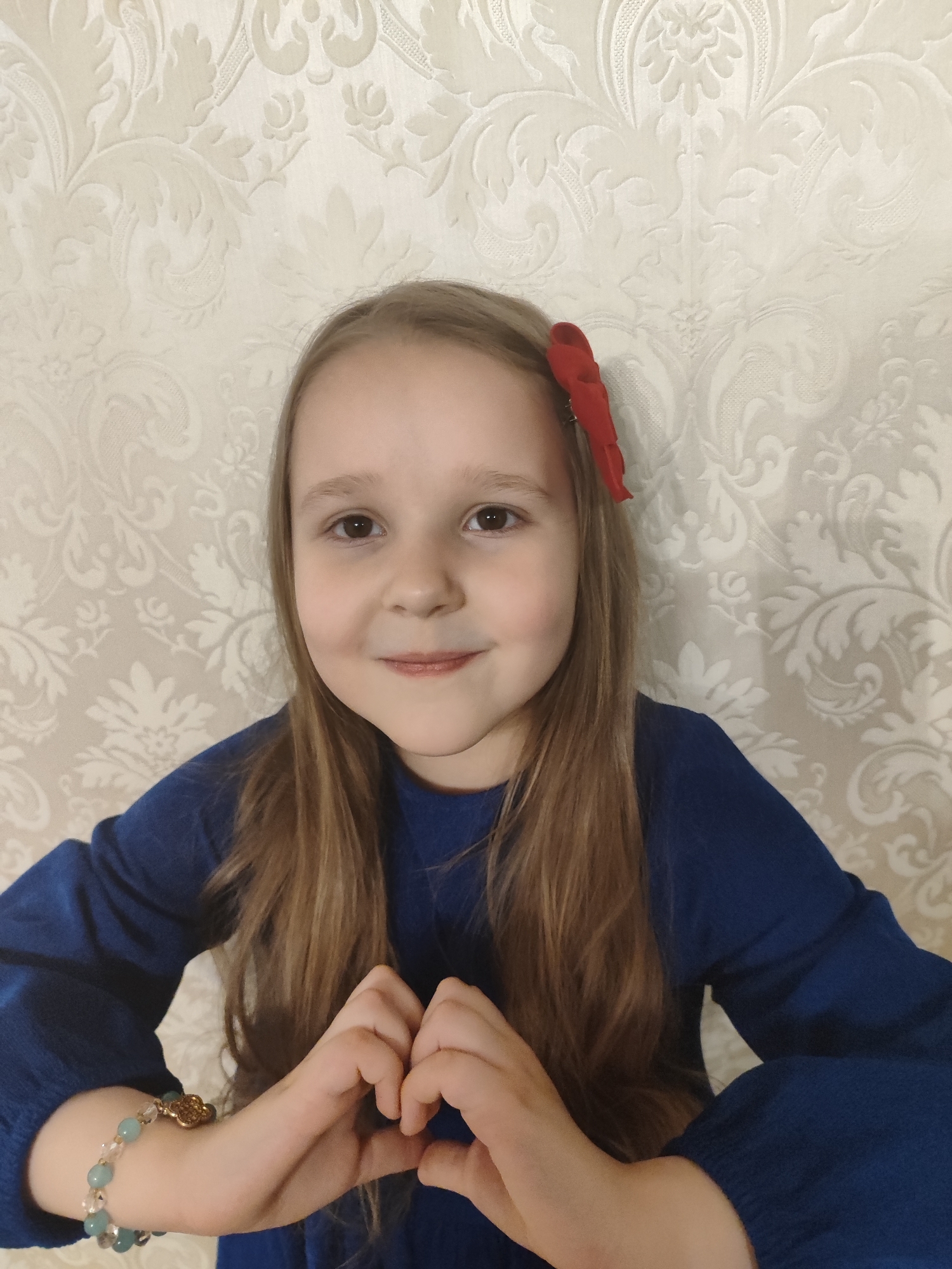 A young girl with long brown hair and a red bow in her hair is centered in the frame. She is wearing a blue long-sleeved shirt. She is looking directly at the camera with a slight smile. Her hands are positioned in front of her, appearing to form a heart shape. A beaded bracelet is visible on her left wrist. The background is a light-colored wall with a floral pattern. There is no indication of the location being Krasnodar, Russia.