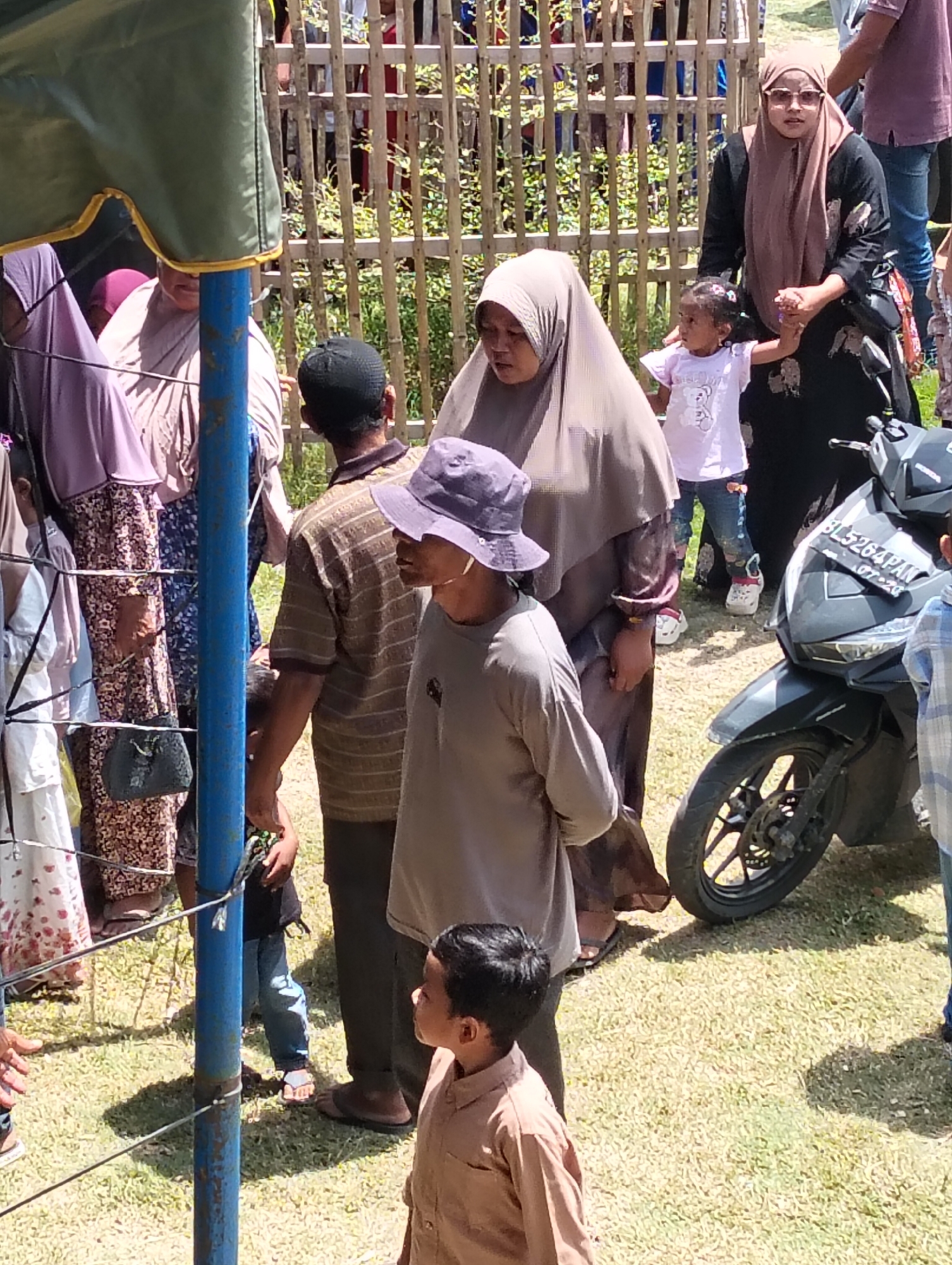 In Grong-Grong, Indonesia, a group of people are gathered outdoors, possibly for an event. A blue pole stands in the foreground, partially obscuring a woman wearing a brown hijab and sunglasses, who is holding the hand of a young child in a white t-shirt. To their left, other individuals in traditional attire are visible. In the background, a wooden fence runs horizontally across the frame, with more people standing behind it. A motorcycle is parked to the right, with its license plate reading "BL 5264 PAN". The ground is covered in dry grass, and shadows suggest a bright, sunny day.