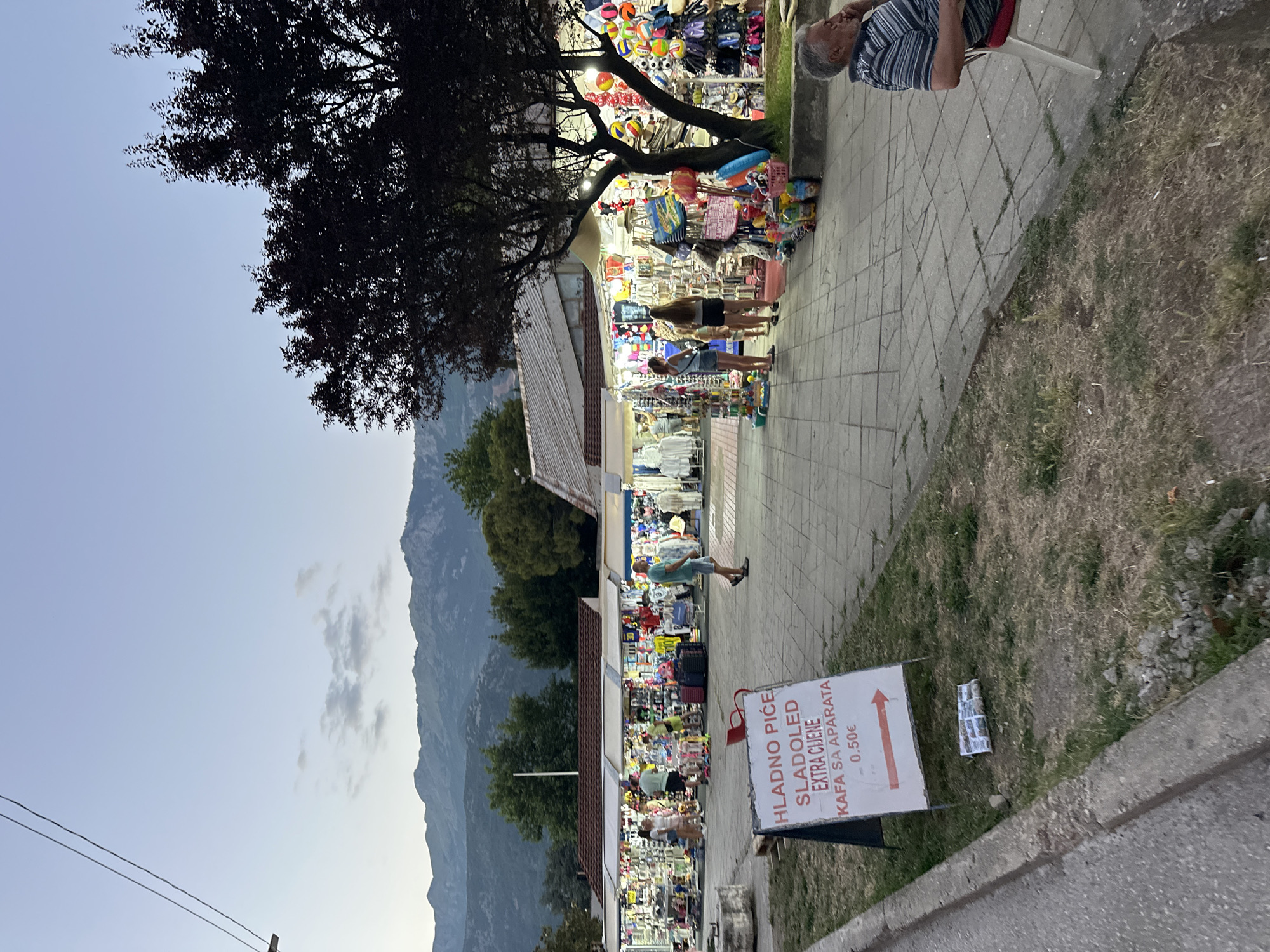 The image depicts an outdoor marketplace scene at dusk, likely in Budva, Montenegro, characterized by a row of stalls selling various goods, including clothing, souvenirs, and beach accessories. In the foreground, a sign in a foreign language advertises "HLADNO PIĆE, SLADOLED, EXTRA CIJENE, KAFA SA APARATA" with a price of "0.50€," indicating beverages, ice cream, good prices, and coffee from a machine. A man is seated on a white chair to the right, looking down and appearing to be resting or contemplating. In the mid-ground, several people are browsing the stalls, and a few are walking along the paved pathway. The stalls are brightly lit, illuminating a variety of merchandise. Behind the stalls, lush green trees are visible, and in the background, majestic mountains rise against a pale twilight sky. The ground is paved with square tiles, and a patch of grass with some weeds runs along the left side of the image. The overall atmosphere suggests a lively but winding-down evening in a tourist-oriented area.