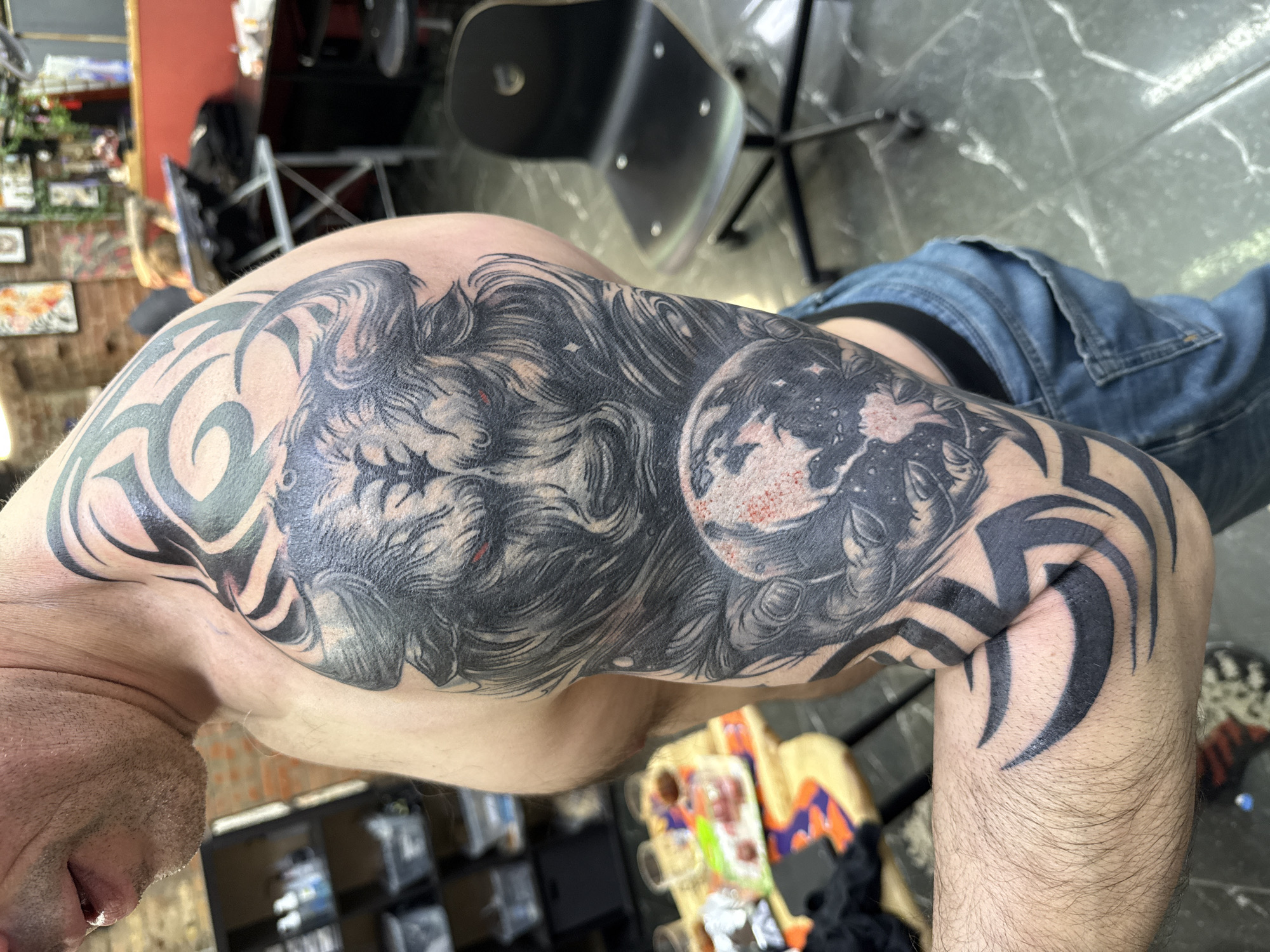 A close-up shot depicts a man's tattooed arm, with the focus on the detailed artwork. The tattoo features a combination of tribal designs, a wolf's head with prominent eyes, and a rendition of the Earth. The man is wearing denim jeans. The setting appears to be indoors, with a black chair and a patterned floor visible in the background. It is not possible to identify the location in the city of Moscow, Russia from the visual information alone.