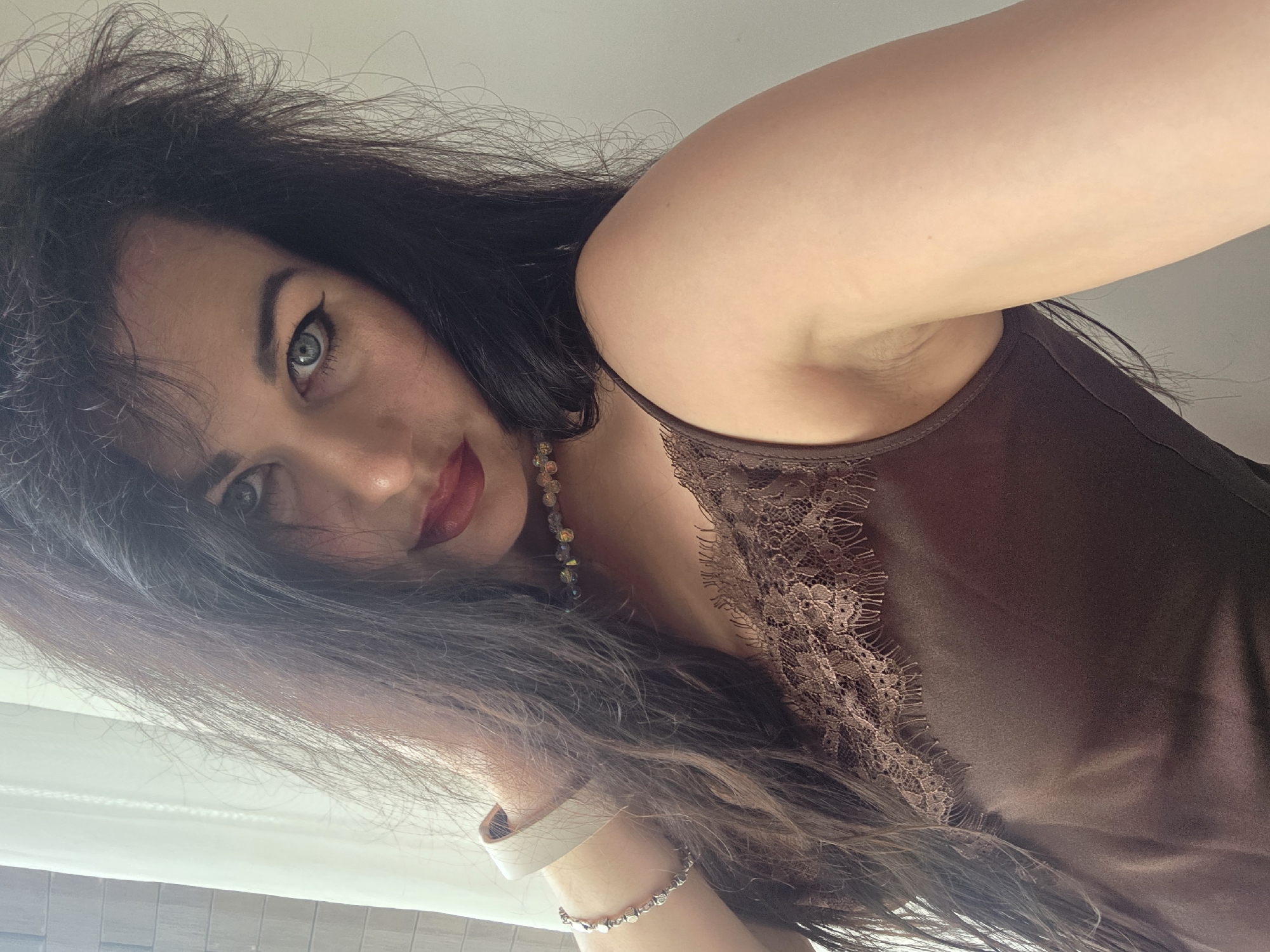 The image is a close-up, chest-up portrait of an adult female in an indoor setting. The individual has long, dark brown to black hair, which appears voluminous and somewhat tousled, framing her face. Her eyes are light blue, accentuated with dark eyeliner and mascara, and she wears deep red lipstick. She is looking directly into the camera with a direct gaze.
She is dressed in a dark brown camisole or slip-style top with a satin-like sheen, featuring intricate lace trim along the neckline. Adorning her neck is a choker-style necklace composed of faceted, multicolored beads that reflect light. On her left wrist, a metallic, possibly silver-toned, chain or linked bracelet is visible, along with a wide-banded watch, likely white or light-colored, with a metallic casing. Her left arm is raised, with the hand out of frame, bringing her shoulder and arm prominently into the shot.
The background on the left side reveals a section of a white curtain or sheer blind, indicating a window, and a portion of a tiled wall with light-colored, rectangular tiles. The right side of the background consists of a plain, light-colored (off-white or light gray) wall. Natural light appears to be illuminating the scene from the left.
Regarding the specified location, the visual content of the image does not contain any discernible elements or landmarks that confirm it was taken in Sochi, Russia.
