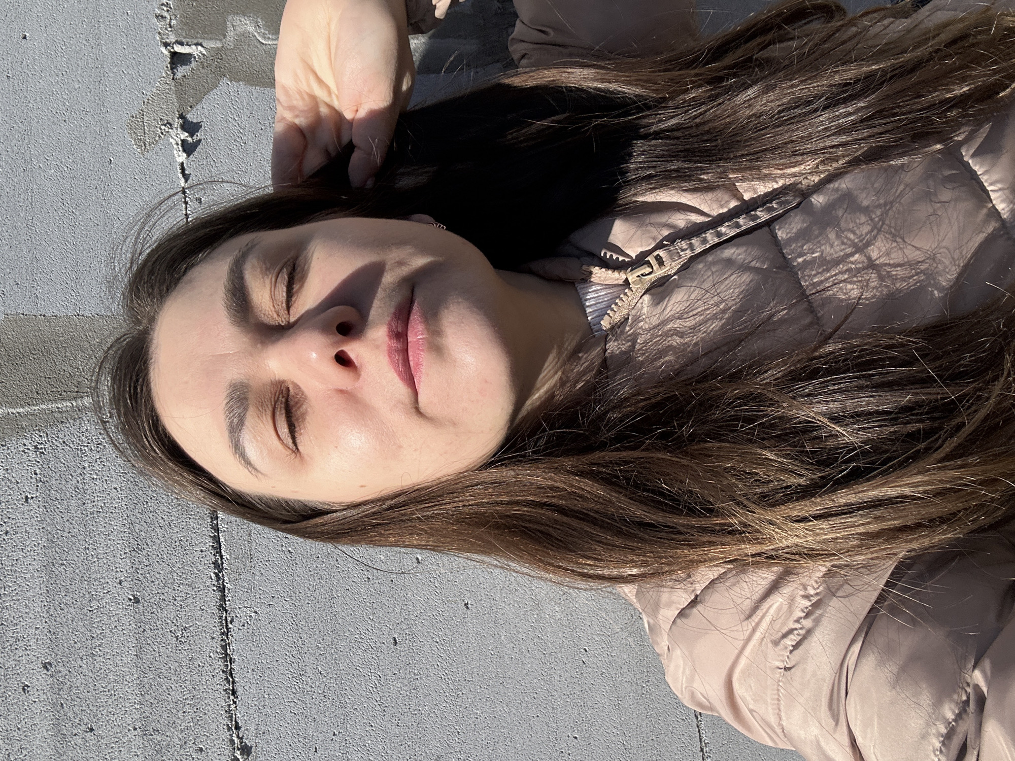 The image depicts a woman lying on a concrete surface, likely outdoors. Her eyes are closed, and her face is turned towards the light. She is wearing a beige puffer jacket. Her long brown hair is spread around her head on the concrete. Her hand is resting near her head. The background appears to be a concrete wall or ground. The location is not identifiable based solely on the image.