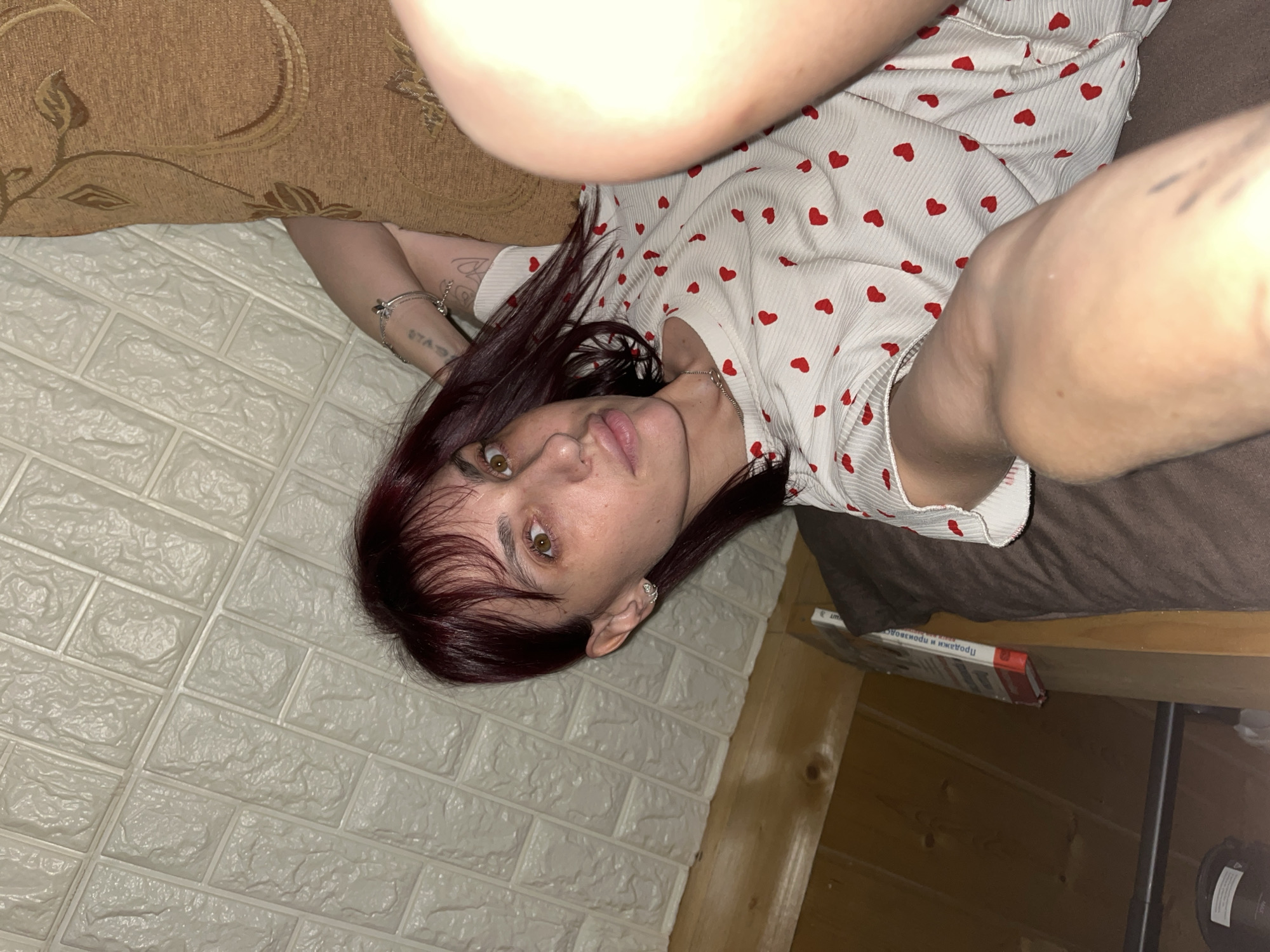 In a room with a textured white ceiling, a person with dark red hair and brown eyes lies on a dark brown surface, propped against a brown cushioned furniture item with a patterned fabric. The person is wearing a white top with a pattern of small red hearts. Their left arm is bent, with tattoos visible on the forearm. The right side of the image is largely obscured by the person's leg, which is positioned towards the camera. The background consists of wooden paneling and a shelf with a book. The overall lighting suggests an indoor environment, possibly at night. There are no identifiable landmarks or specific details to place this scene within the city of Нагорный, Russia.