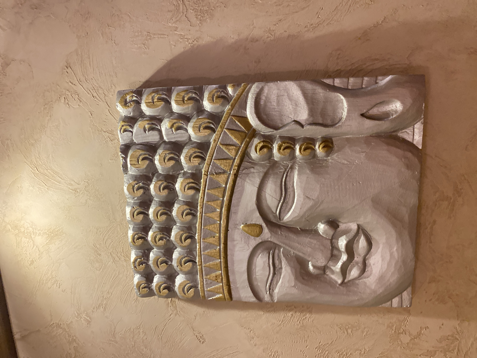 This media file depicts a decorative relief sculpture of a face, likely a deity or a stylized portrait, mounted on a textured wall. The sculpture is primarily silver with accents of gold. The face is rendered in profile, with prominent features such as closed eyes, a curved nose, and full lips. The left side of the sculpture features a repeating pattern of curved elements, each outlined in gold, resembling scales or stylized curls. A decorative border with triangular gold patterns separates this patterned section from the facial features. The background wall has a rough, stucco-like texture and is illuminated by a warm light source, casting a subtle shadow from the sculpture onto the wall to the upper left. The overall impression is one of artistic craftsmanship and a serene, perhaps spiritual, aesthetic. There is no visible text in the image.