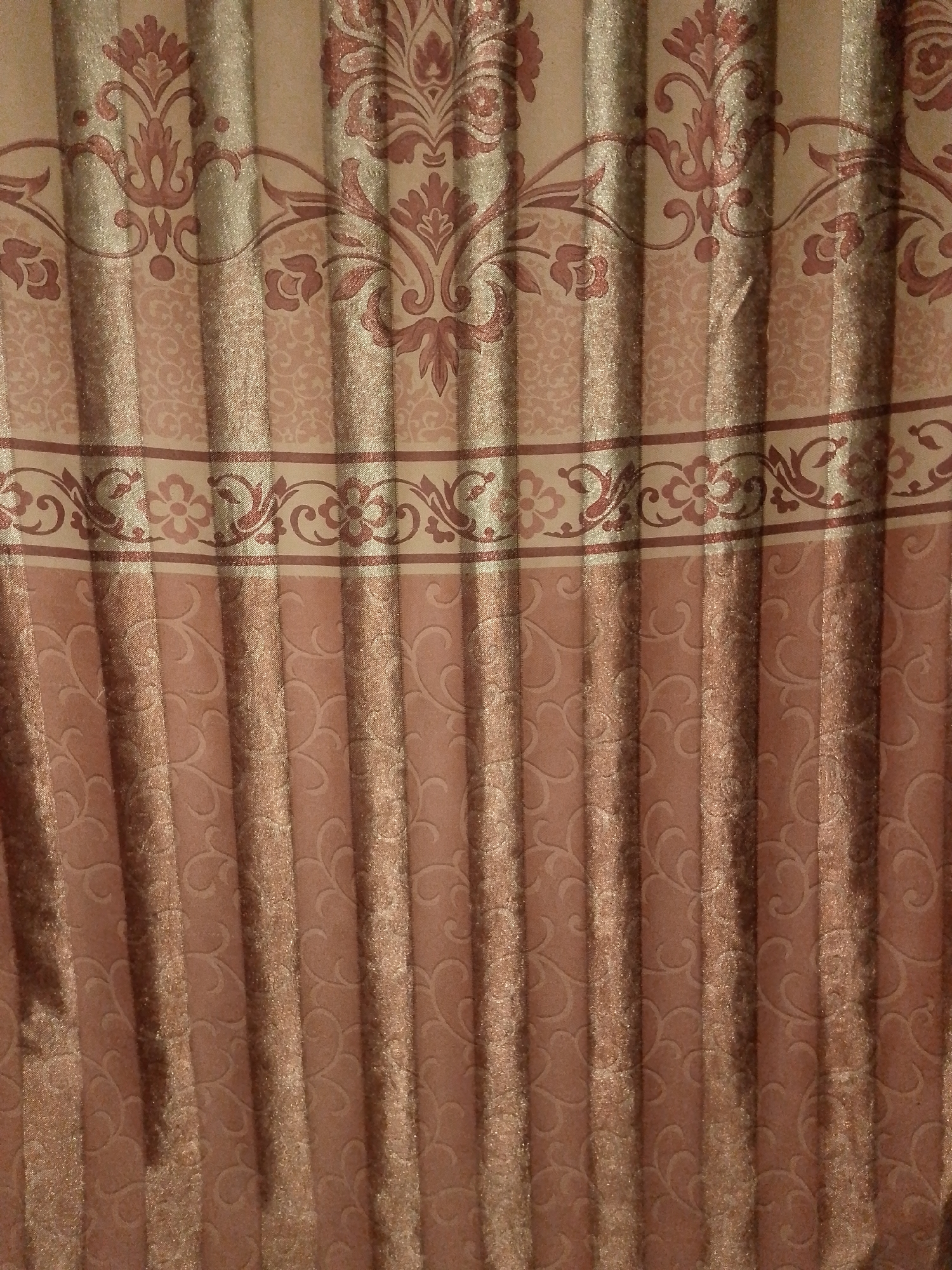 A close-up, full-frame shot displays vertically pleated drapes with a damask pattern. The drapes are primarily a dusty rose color with metallic gold threads woven into them, giving them a subtle shimmer. A wide band near the top features a more intricate damask pattern in a darker rose hue against the gold fabric. Below this band, a narrower border showcases a repeating floral and vine motif in the same darker rose. The bottom half of the drapes features a more subtle, embossed damask pattern in the dusty rose color. The lighting creates strong vertical shadows along the pleats, highlighting the texture and sheen of the fabric. No people or specific locations within Abuja, Nigeria, are visible in this image; it solely focuses on the textile itself.