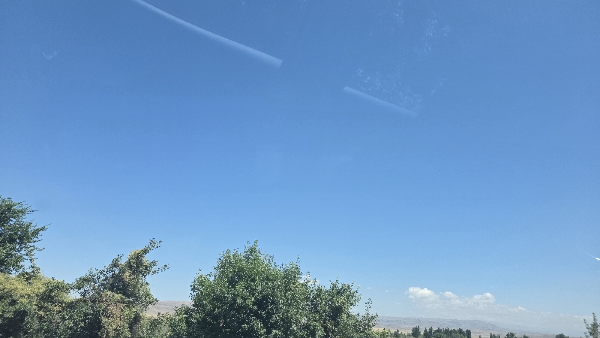 The image captures a clear blue sky with some faint reflections visible, possibly from a window. Below the sky, lush green trees are visible in the foreground, with their branches reaching upwards. In the distance, a slightly hazy landscape of rolling hills or mountains can be seen. The overall scene suggests a bright, sunny day in a natural setting, potentially on the outskirts of Birdik, Kyrgyzstan.
