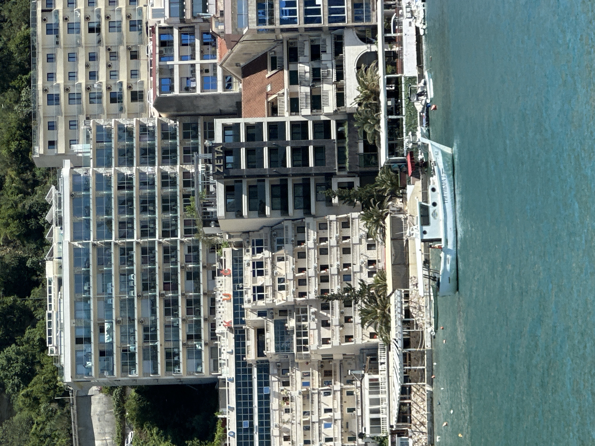 The image shows an aerial view of Rafailovići, Montenegro, a coastal town, highlighting the intersection of architecture and the Adriatic Sea. Buildings of varying heights and designs stand close together, with many featuring balconies and numerous windows. A modern, dark-colored building labeled "ZETA" is visible. The buildings are made of light-colored materials. Along the shoreline, a boat is docked, and the clear turquoise water of the sea is a dominant visual element. There are trees alongside the buildings. People can be seen near the boat.