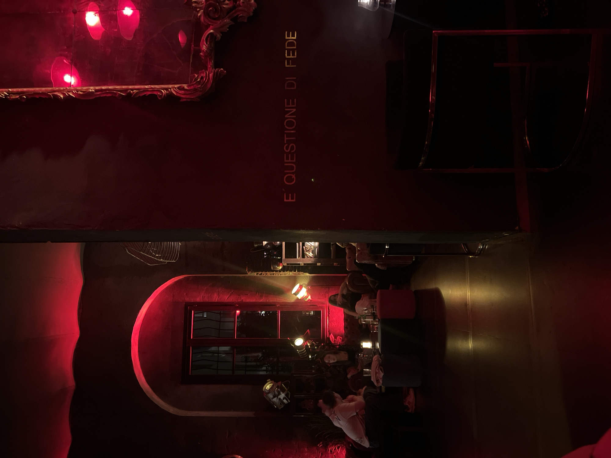 The image captures the dimly lit interior of a bar or lounge, exuding a moody and intimate atmosphere primarily cast in red light, suggesting it's nighttime or evening.
On the left side of the scene, a dark wall displays an ornate, gilded mirror frame near the top. Below the mirror, golden text is vertically inscribed on the wall, reading "E' QUESTIONE DI FEDE" (Italian for "It's a matter of faith"). The wall is a deep maroon or dark red hue.
Towards the right side of the frame, several patrons are visible, seated at tables and engaged in socializing. At least three to four individuals can be discerned. They are surrounded by dark furniture, including chairs and what appear to be red stools or small sofas. Various decorative lamps and spotlights provide localized yellowish light in this area, illuminating parts of the dark, reflective floor.
Architectural details include an arched window with dark panes, set into the red-lit wall, and industrial-style lighting fixtures hanging or mounted throughout the space. A unique decorative object resembling a helmet or mask is visible on a table in the foreground on the right. The overall impression is of a stylish establishment in Milan, Italy, designed for leisure and social gatherings.