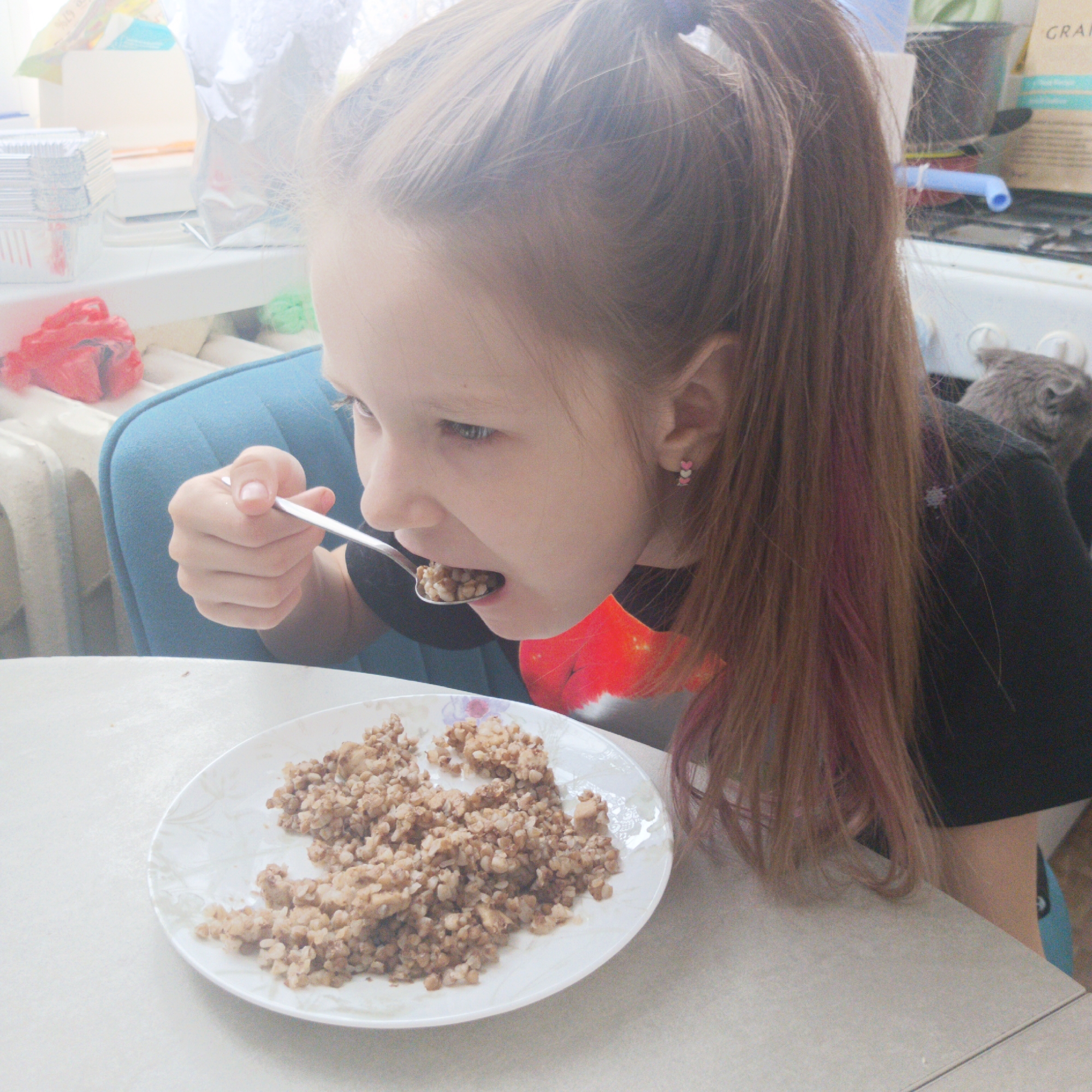 A young girl with reddish-blonde hair pulled back in a ponytail is eating buckwheat from a white plate with a spoon. She is wearing a black shirt with a red design. She is sitting at a light-colored table with a blue chair behind her. In the background, there is a window with some items on the windowsill, a radiator, and part of a stove with a gray cat sitting on or near it. Based on the location identified in the prompt, this image was likely taken in Chaikovsky, Russia.