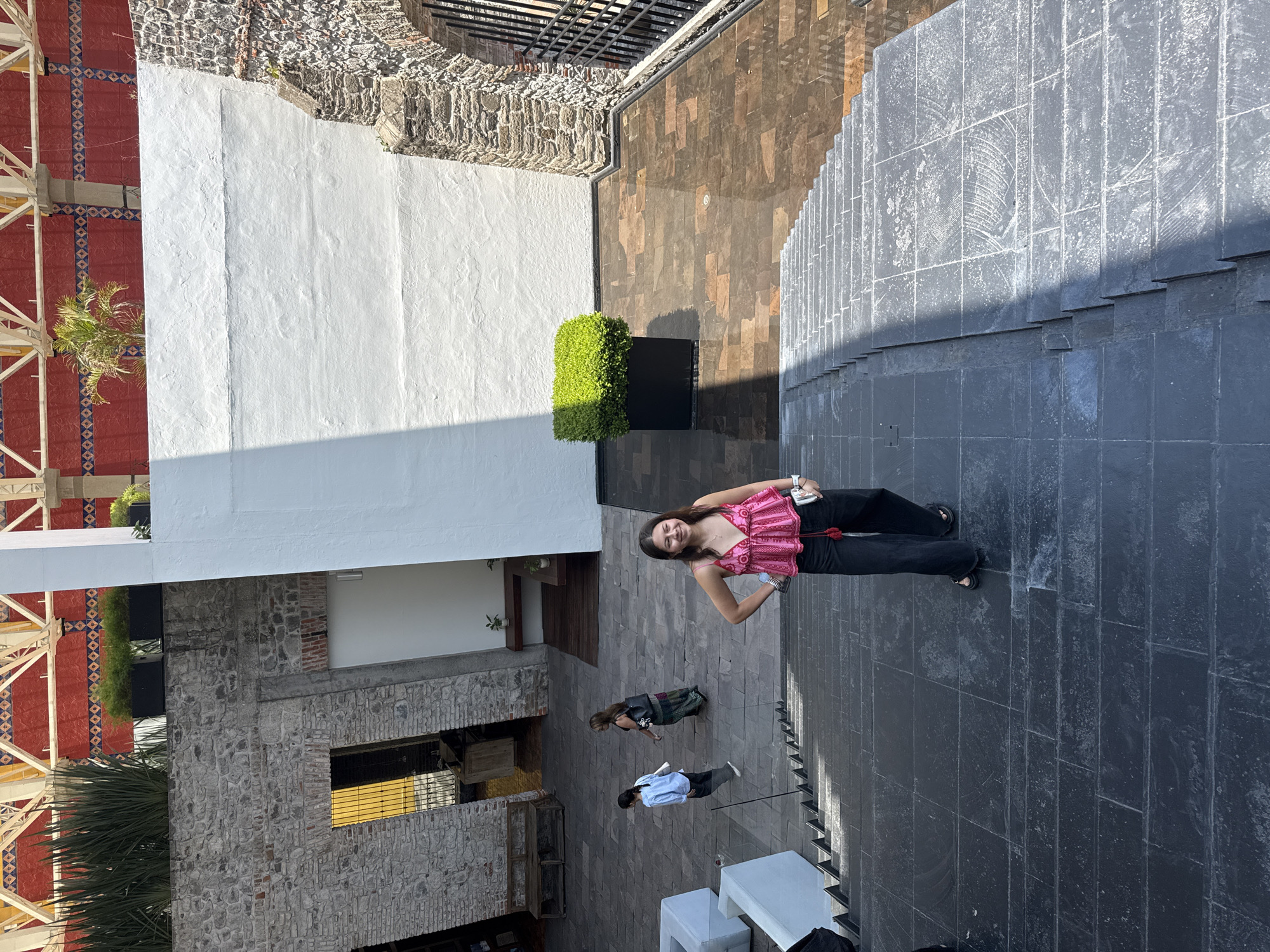 This image captures an outdoor architectural space from an elevated perspective, looking down into a courtyard or multi-level area.
In the foreground, a young woman stands on a dark grey tiled floor, smiling brightly and looking up towards the camera. She wears a pink, intricately patterned sleeveless top, black trousers, and dark sandals. A watch is visible on her left wrist, and she holds a small item, possibly a phone or camera, while a small red bag hangs from her waist.
Further back on the same dark tiled surface, two other individuals are present. One person, dressed in a dark top and a patterned skirt, stands a short distance behind the main subject. Another person, wearing a light blue shirt and dark pants, is seated or leaning on white outdoor furniture located even further back.
The surrounding architecture features a blend of materials and styles. The ground is predominantly covered in large, rectangular dark grey tiles, which transition into a set of descending steps on the right side of the frame. To the left of the main subject, a planter with a vibrant green, manicured shrub is visible, followed by a section of floor made of irregular brown and earthy-toned tiles. Beyond these, a stark white stucco wall rises, flanked by a wall constructed from rough-hewn stone. On the far left, a distinct red tiled area with blue and white geometric patterns is prominent, supported by white metal structures, suggestive of a traditional Mexican design.
The scene is well-lit by bright daylight, casting defined shadows, indicating a clear, sunny day. The architectural elements, particularly the stone walls and patterned tiles, provide strong contextual cues for Puebla, Mexico. No visible text is present in the image. The overall atmosphere, influenced by the main subject's smile, appears cheerful and relaxed.