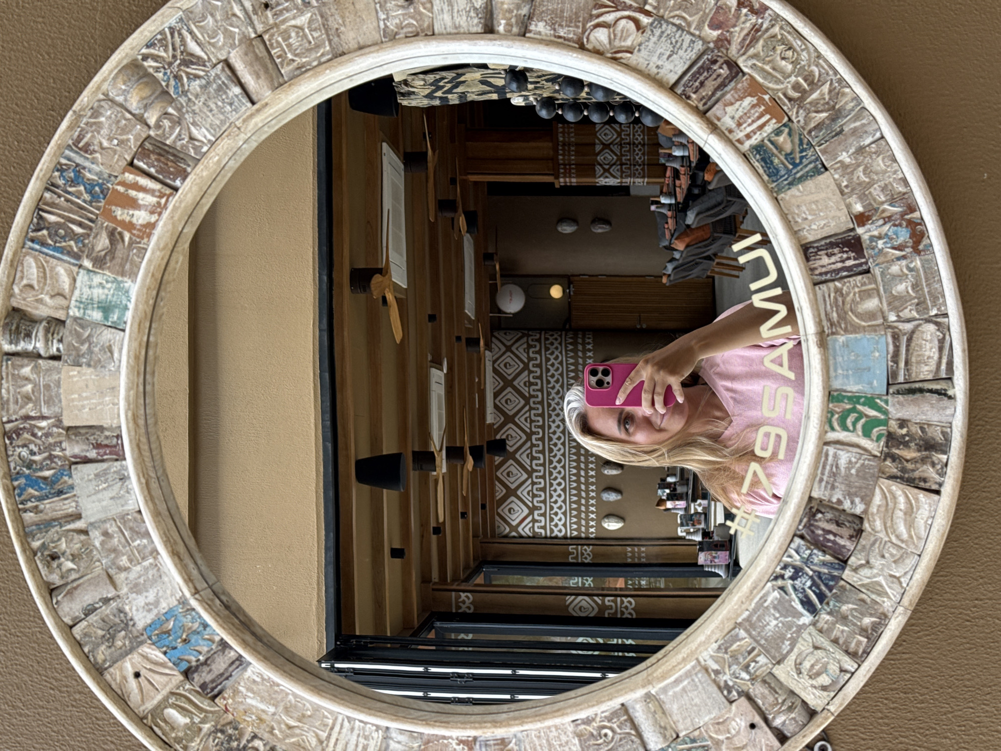 A woman is taking a mirror selfie in what appears to be a restaurant in Ko Samui District, Thailand. The mirror is round with an ornate, tiled frame. The woman has blonde hair and is wearing a pink t-shirt. She is holding up a pink smartphone to take the photo. In the reflection of the mirror, tables and chairs are visible, suggesting a dining area. The restaurant has a rustic interior with wooden accents and decorative wall art. The woman's t-shirt has a text overlay that reads "#79SAMUI".