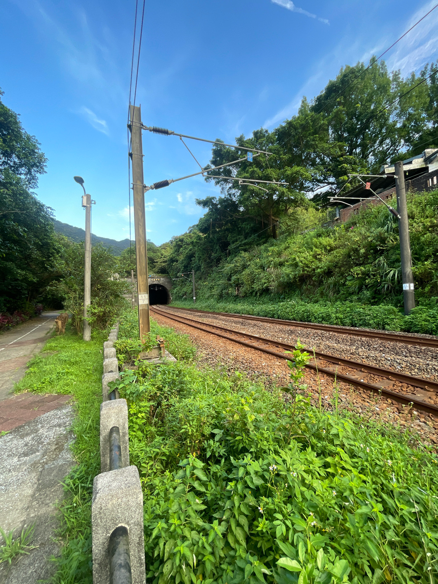 An outdoor shot shows a railway tunnel entrance in New Taipei City, Taiwan, on a clear day. The tunnel is arched and made of stone. A double track railway line leads into the tunnel, with ballast and gravel visible on the tracks. To the left of the tracks is a paved pathway and a concrete barrier with metal railings. A tall wooden utility pole with overhead wires stands to the right of the pathway, and another similar pole is located closer to the railway tracks. Green trees and vegetation cover the surrounding hillsides, with a blue sky and scattered white clouds overhead. A street lamp is visible on a pole to the left of the pathway.