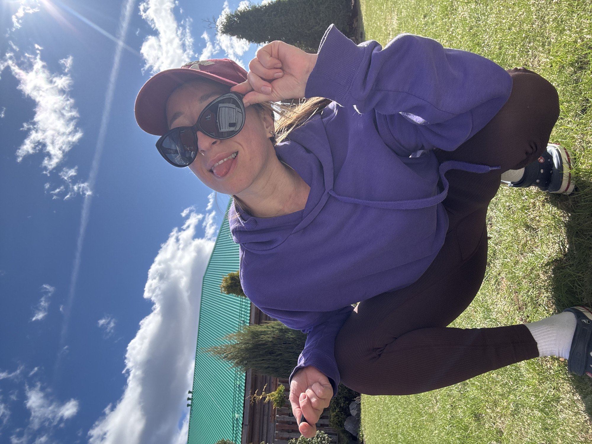 A woman squats outdoors on grass. She wears a purple hooded sweatshirt, brown leggings, a dark red baseball cap, and black sunglasses. Her tongue is slightly extended. She appears to be on green grass in what seems to be a backyard setting. A green-roofed building and blue sky with white clouds are visible in the background. A partial view of a fence and greenery are at the bottom of the image. The location of the photo is in Прикамский, Russia.