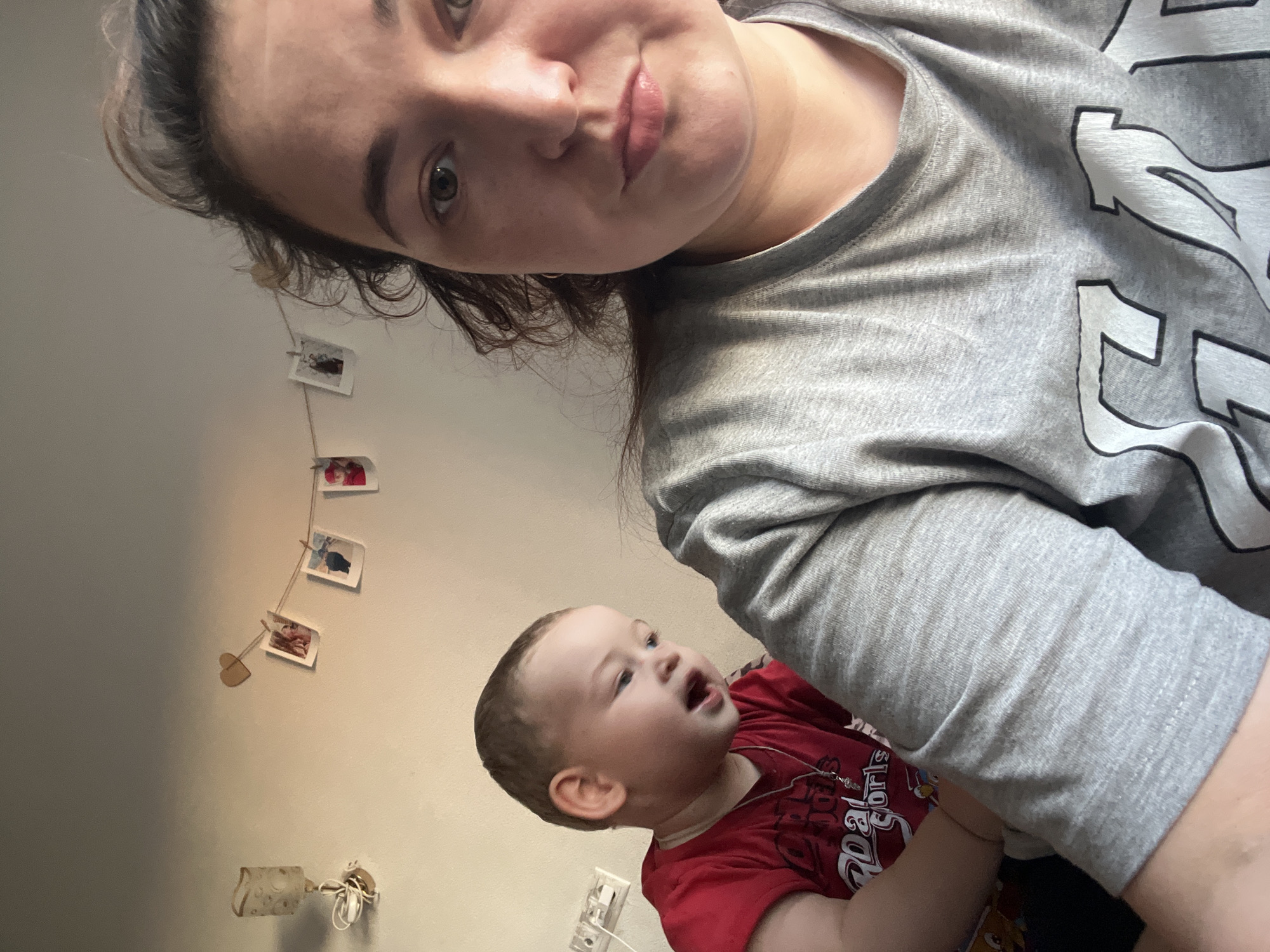 The media file is a close-up, low-angle selfie of two people. In the foreground, a woman with dark hair and light eyes is looking directly at the camera. She is wearing a grey t-shirt. To her right and slightly behind her, a baby with light skin and brown hair is looking up and to the right with an open mouth, as if speaking or surprised. The baby is wearing a red t-shirt. The background is a plain wall with a string of small photos hanging on it, secured by clothespins. There is also a light fixture attached to the wall. The lighting suggests it is daytime. The overall mood appears casual and intimate, capturing a candid moment between the woman and the baby.
