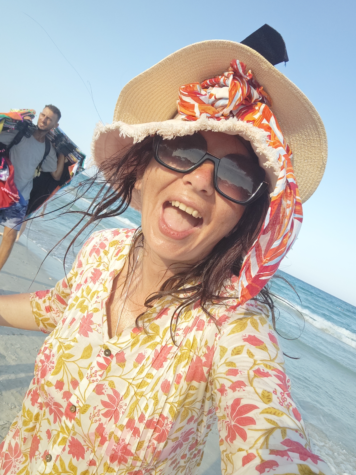 A woman with dark, wet hair is taking a selfie on a beach in Mahdia, Tunisia. She is wearing a wide-brimmed straw hat adorned with a colorful scarf, sunglasses, and a white blouse with a floral print in pink and yellow. Her mouth is open, and her tongue is visible. Behind her, a man with dark hair and a beard is walking away from the camera, carrying a large bundle of colorful items on his shoulders, possibly merchandise. The ocean with moderate waves is visible in the background, and the sky is clear and blue. The ground is sandy.