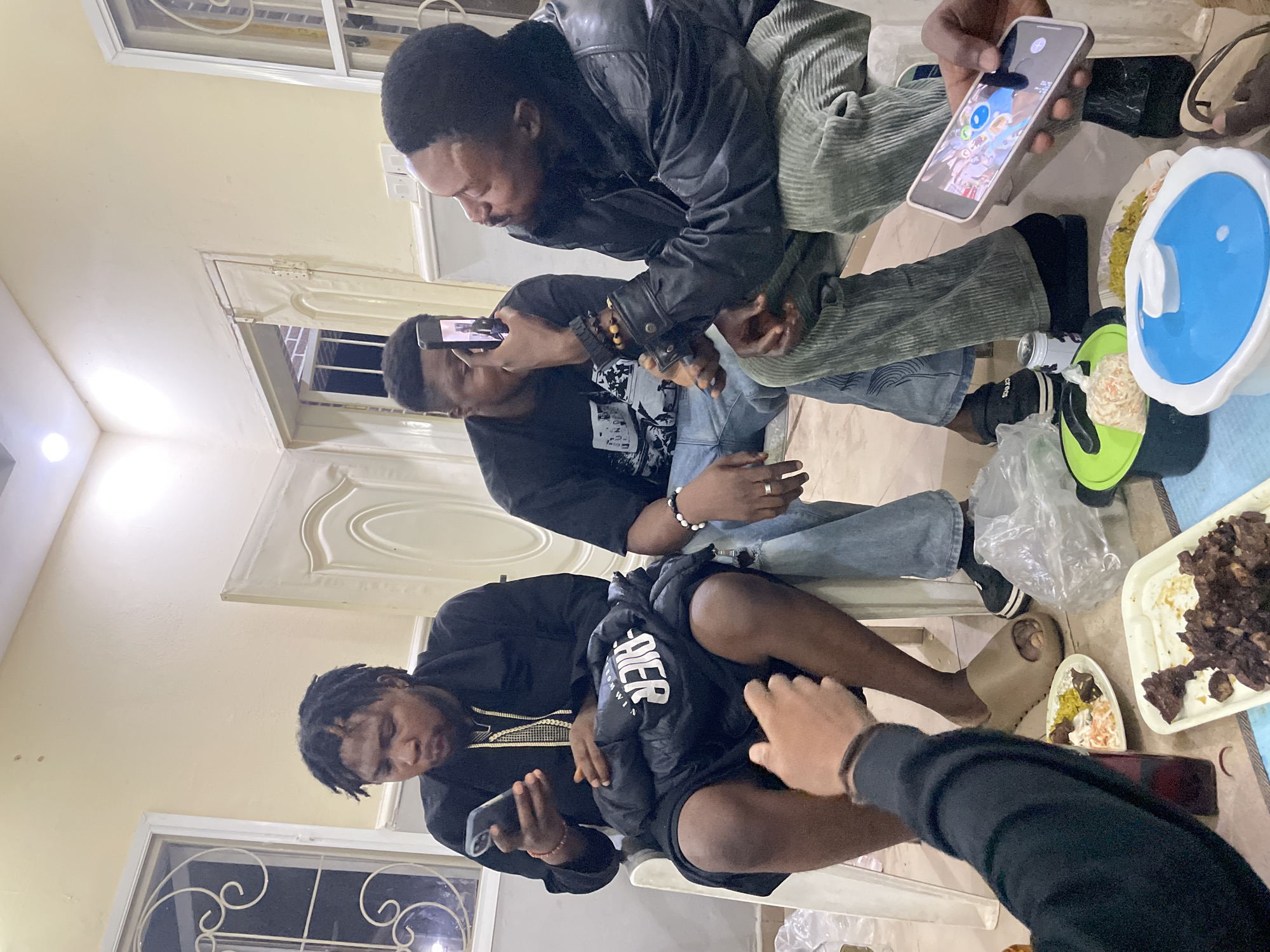 In an indoor setting, possibly a residence in Jos, Nigeria, four individuals are engaged with their mobile phones. Three of them are seated on white plastic chairs. One person wears a black leather jacket and green corduroy pants. Another wears a black shirt and blue jeans. A third wears a black jacket and black shorts. A fourth person is partially visible on the bottom right. Food containers, including a blue lidded container, a green dish, and trays with food items, are scattered on the floor around them. A clear plastic bag is also visible. The environment is lit with overhead lighting.