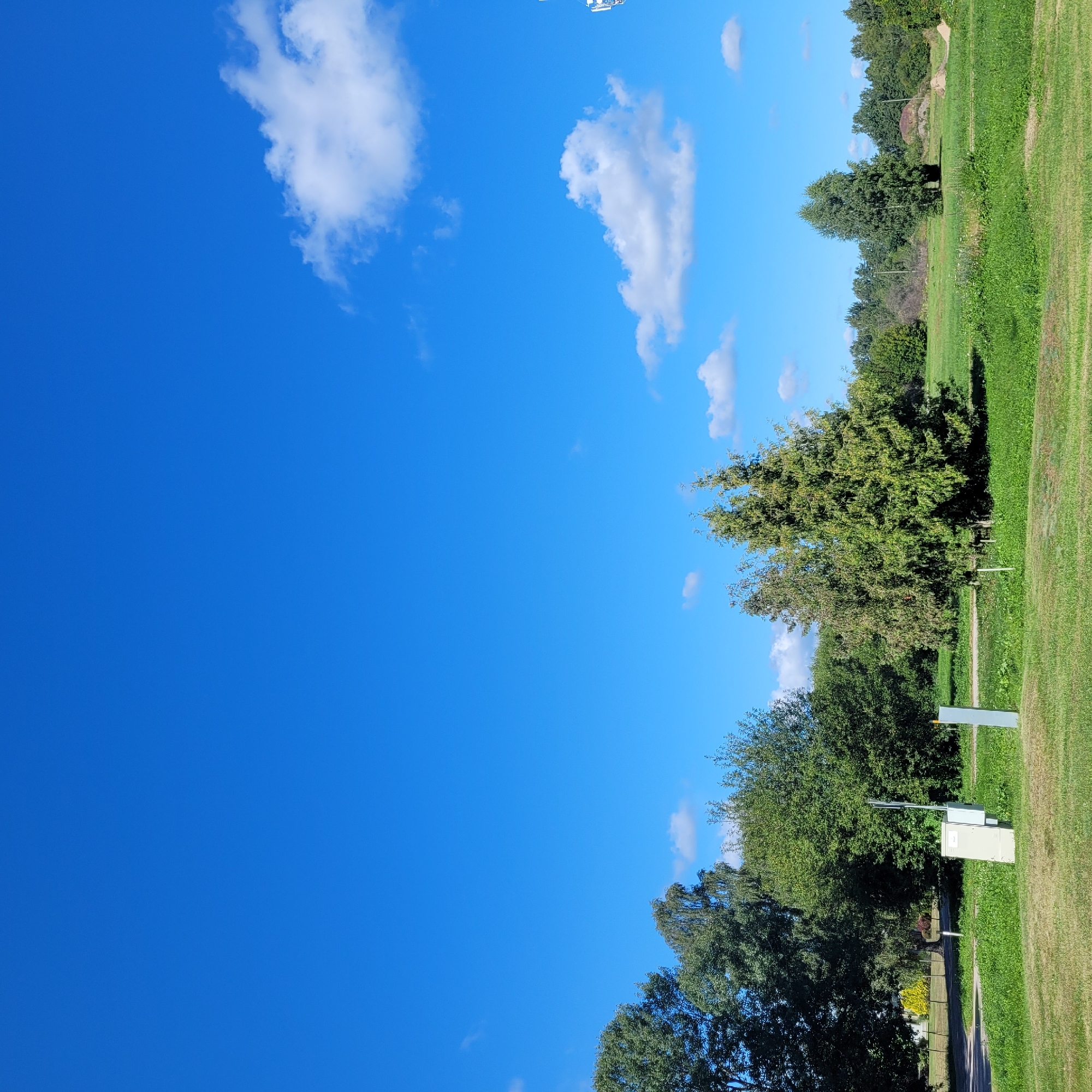 This is a wide, ground-level outdoor shot of a park or open grassy area under a clear blue sky with scattered white clouds. The foreground is dominated by a well-manicured lawn. In the middle ground, there is a line of trees with varying shades of green foliage, some appearing denser than others. To the left, a paved path is visible, leading into the trees. Several small, light-colored utility boxes or cabinets are situated on the grass near the path, along with a tall, slender grey post. In the distance, more trees form a dense green backdrop, and beyond that, a mound of sand or dirt is visible on the right side of the frame. A very tall, thin white structure, possibly a tower or antenna, is partially visible in the far upper right corner. No people or animals are present in the image.