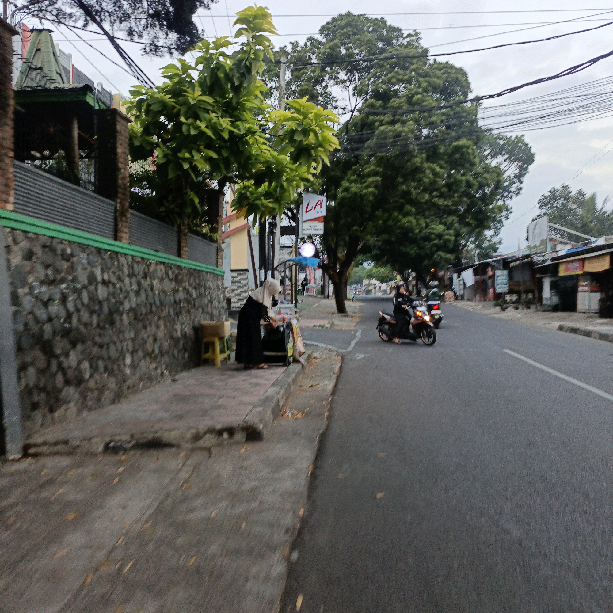 The image depicts a street scene in Salatiga, Indonesia, likely during daylight hours, with an overcast sky. On the left side of the road, a pedestrian walkway is bordered by a stone wall topped with a green trim. A woman wearing a hijab and a long black garment is standing on the walkway, attending to a small mobile cart. Beside her, a yellow stool with a cardboard box on it is visible. A tree with light green leaves is situated behind the woman.
Across the street, a paved road with a white lane marker runs parallel to the walkway. Two motorcycles are on the road. The first motorcycle is being ridden by a person wearing a black outfit and a helmet, moving away from the viewer. A second motorcycle is partially visible behind the first.
In the background, several small buildings with awnings line the right side of the road. Overhead, a complex network of power lines crisscrosses the sky, with trees interspersed throughout the scene. A white sign with red lettering that reads "LA" is visible above the walkway. The overall impression is of a typical urban street in Indonesia.