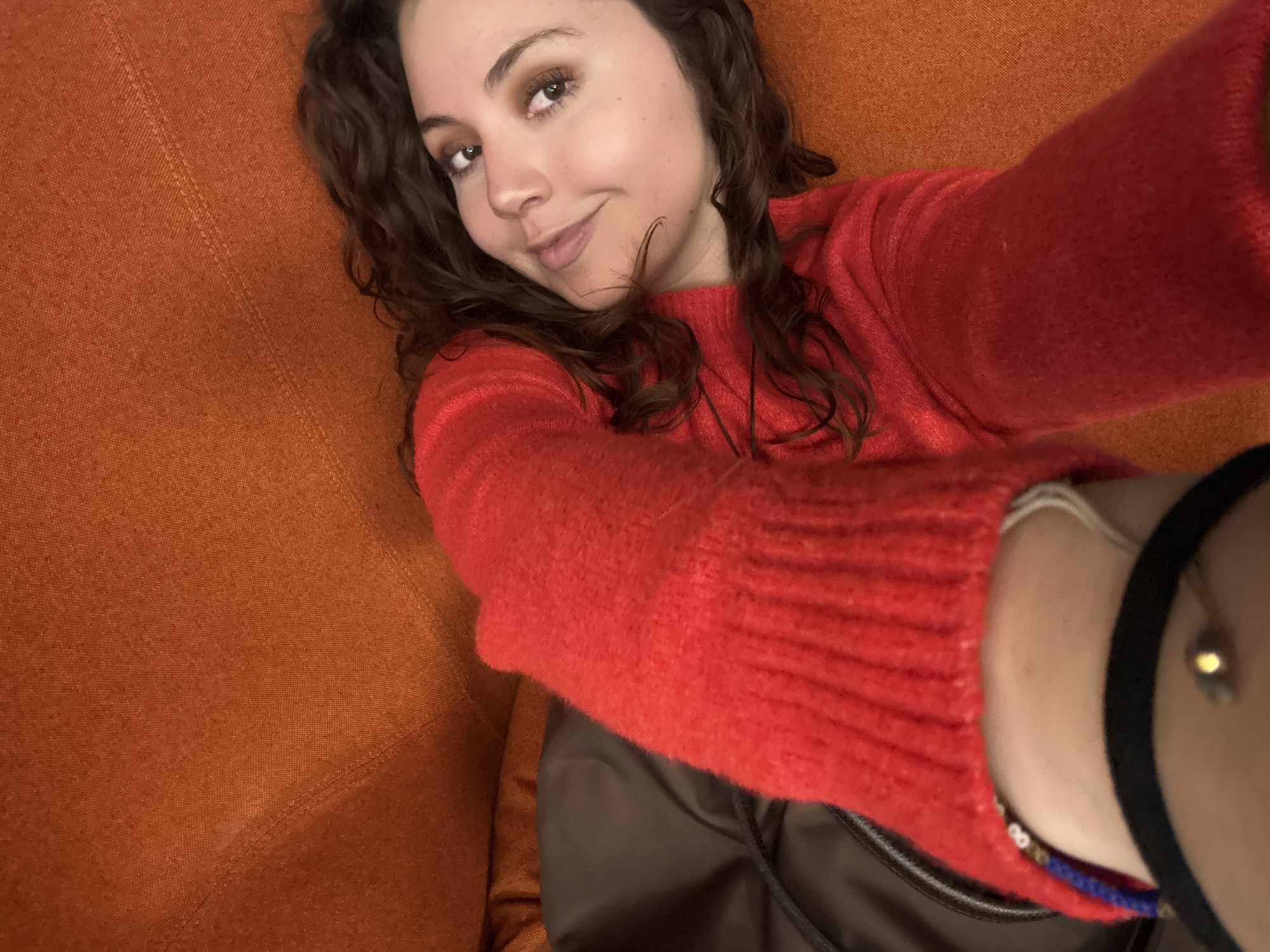 A young woman is taking a selfie indoors. She has curly brown hair, brown eyes, and fair skin. She is wearing a red sweater. She is sitting or lying on an orange couch. Her left arm is extended towards the camera, with bracelets visible on her wrist. A brown bag or jacket is visible near the bottom of the frame. The location is likely Mexico City, Mexico, but there is no verifiable evidence to confirm this assumption from the image.