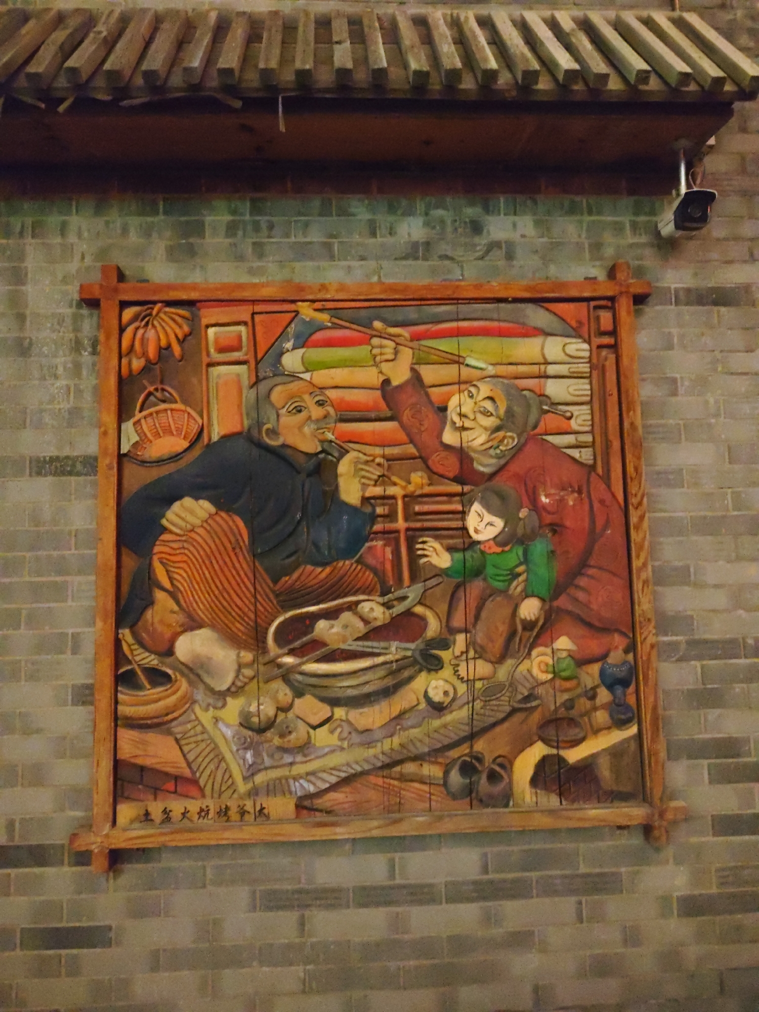 This image depicts a carved wooden mural on a brick wall in Lindian, China. The mural portrays a lively scene of a family, likely preparing food. In the foreground, an elderly man with a dark blue robe and striped orange pants sits, smoking a pipe and looking towards a cooking pot. Beside him, an elderly woman in a red robe is holding a young child wearing a green top and brown pants. The child is reaching out towards the woman. The background features stacked items, possibly food items like corn. The mural is framed by a rustic wooden border. The brick wall shows signs of age and weathering. A security camera is visible mounted on an overhang above the mural. The overall impression is a depiction of traditional rural life and family interaction.