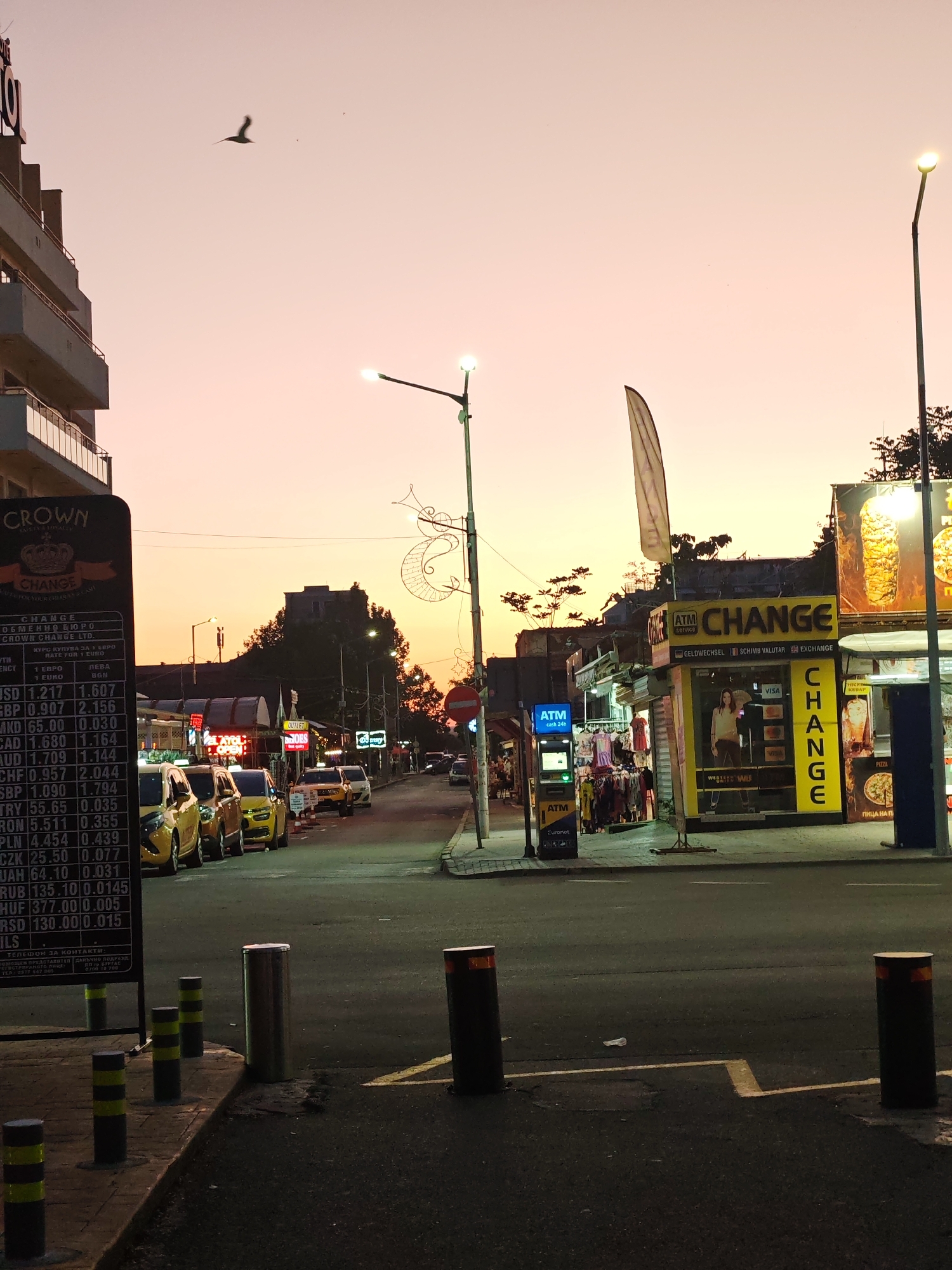 This image depicts a street scene in the city of Nesebar, Bulgaria. The photograph is taken at dusk, showing a street lined with buildings, currency exchange booths and shops. On the left, a sign for "CROWN CHANGE" displays currency exchange rates. Several yellow taxis are parked along the street. Further down the street are more shops and people walking. There are street lights and bollards along the road.