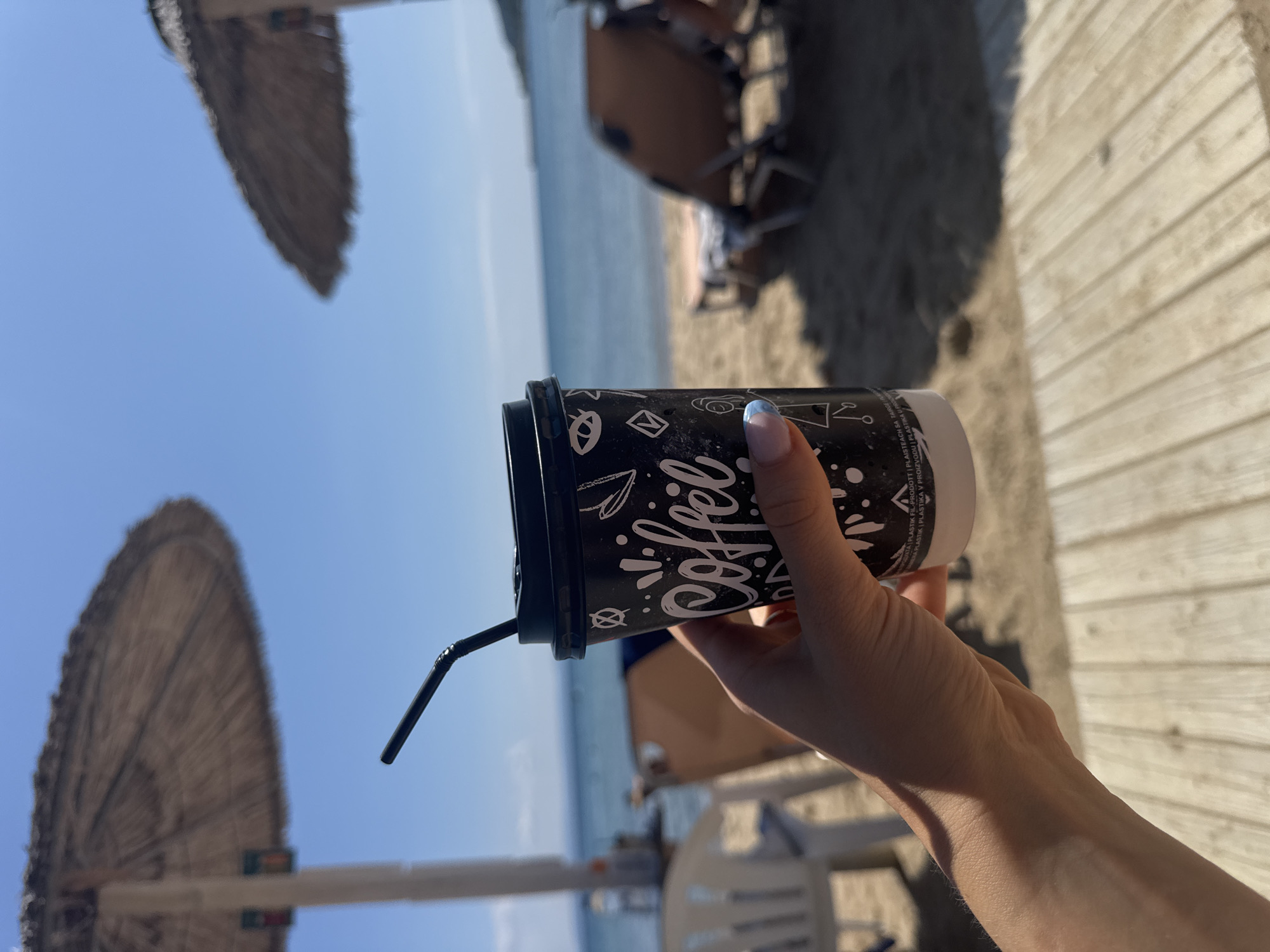 A person's hand, with light blue and pink painted fingernails, holds a black paper coffee cup with white graphics and the word "coffee" written in script. The cup has a black lid and a black straw. The background shows a sandy beach with two thatched umbrellas, lounge chairs, and the blue sea under a clear sky. The scene appears to be a beach in Stavros, Greece, given the context.