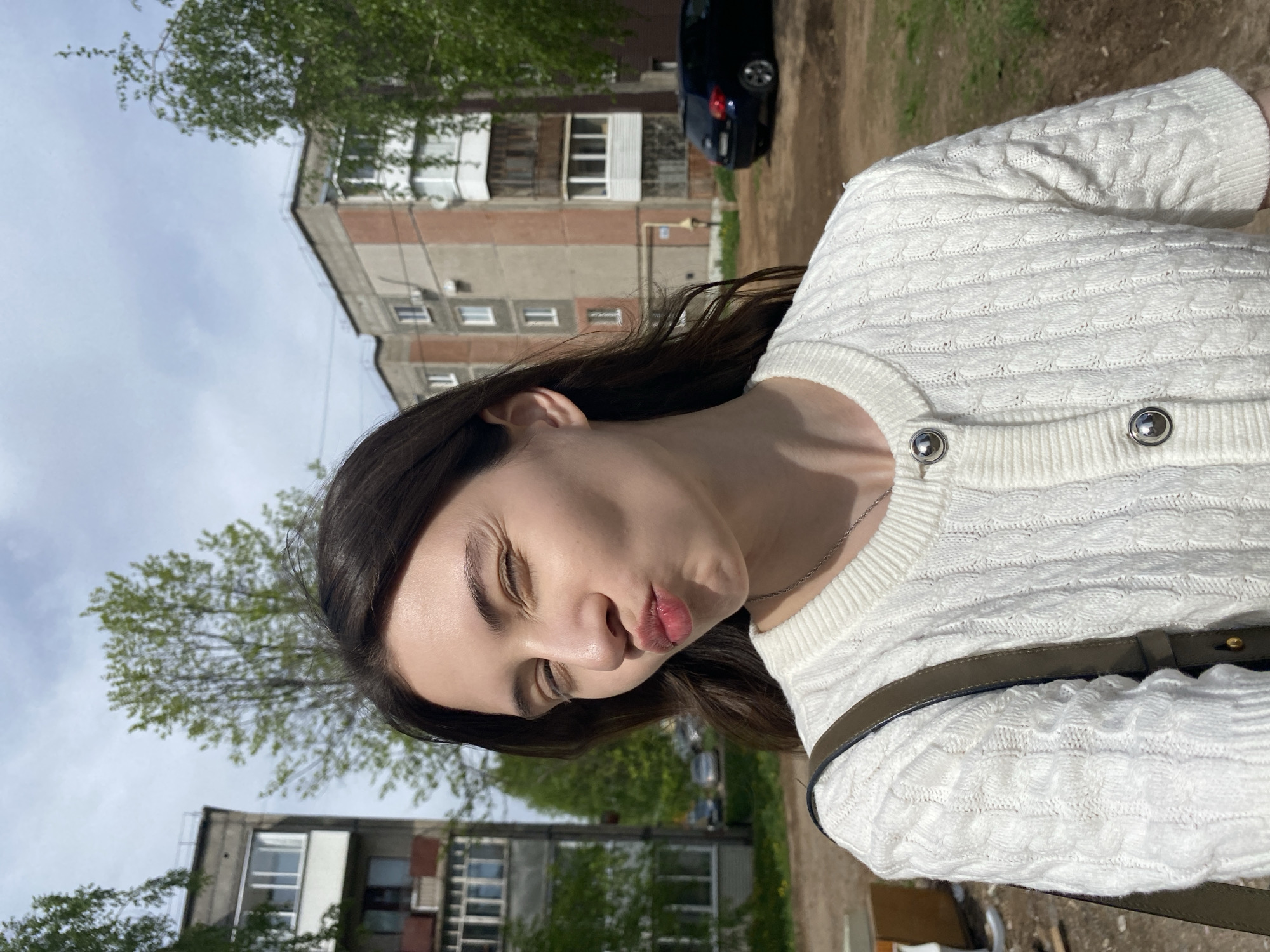 A woman is posing outdoors in the city of Votkinsk, Russia. She has dark hair, wears a white cable-knit sweater, and has a dark-colored bag slung over her shoulder. Her eyes are slightly closed, and she is making a pouting face. The background includes apartment buildings, trees, and a parked black car. The sky is overcast. The setting suggests a residential area on a spring day.