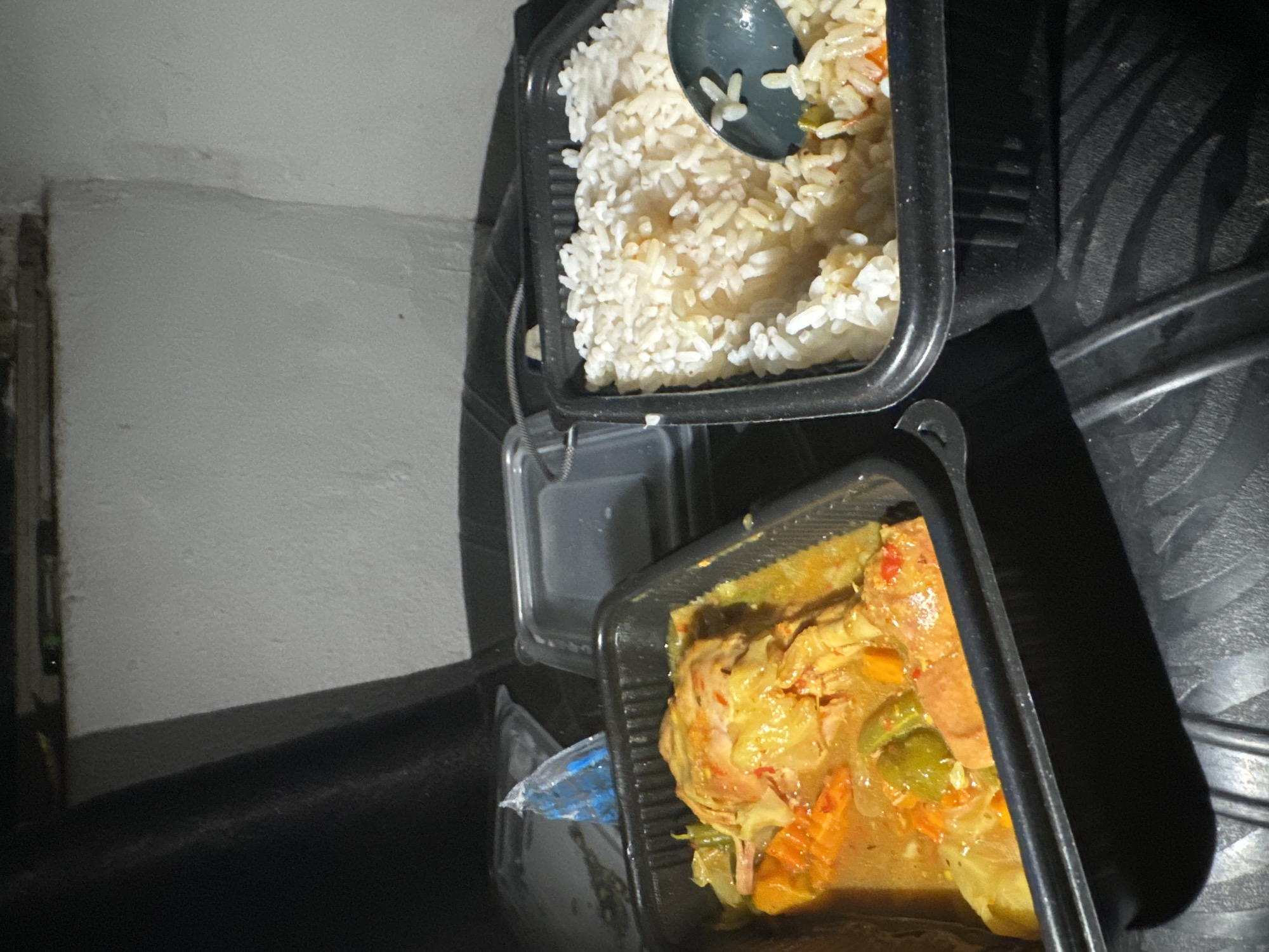 Two black plastic takeout containers of food are arranged on a dark, textured surface, possibly a car's dashboard or trunk liner. The container on the left holds a stew with large pieces of meat, possibly chicken or pork, along with chopped vegetables such as cabbage, carrots, and green peppers. The stew has a reddish-orange hue. The container on the right is filled with cooked white rice, and a dark-colored spoon is partially submerged in it. The background is out of focus, showing a white, textured wall with some dark shadows. The lighting is dim, suggesting an indoor setting or nighttime. No people are visible in the image. This scene is consistent with food prepared for takeout in Awka, Nigeria, where such containers are commonly used for street food and casual dining.