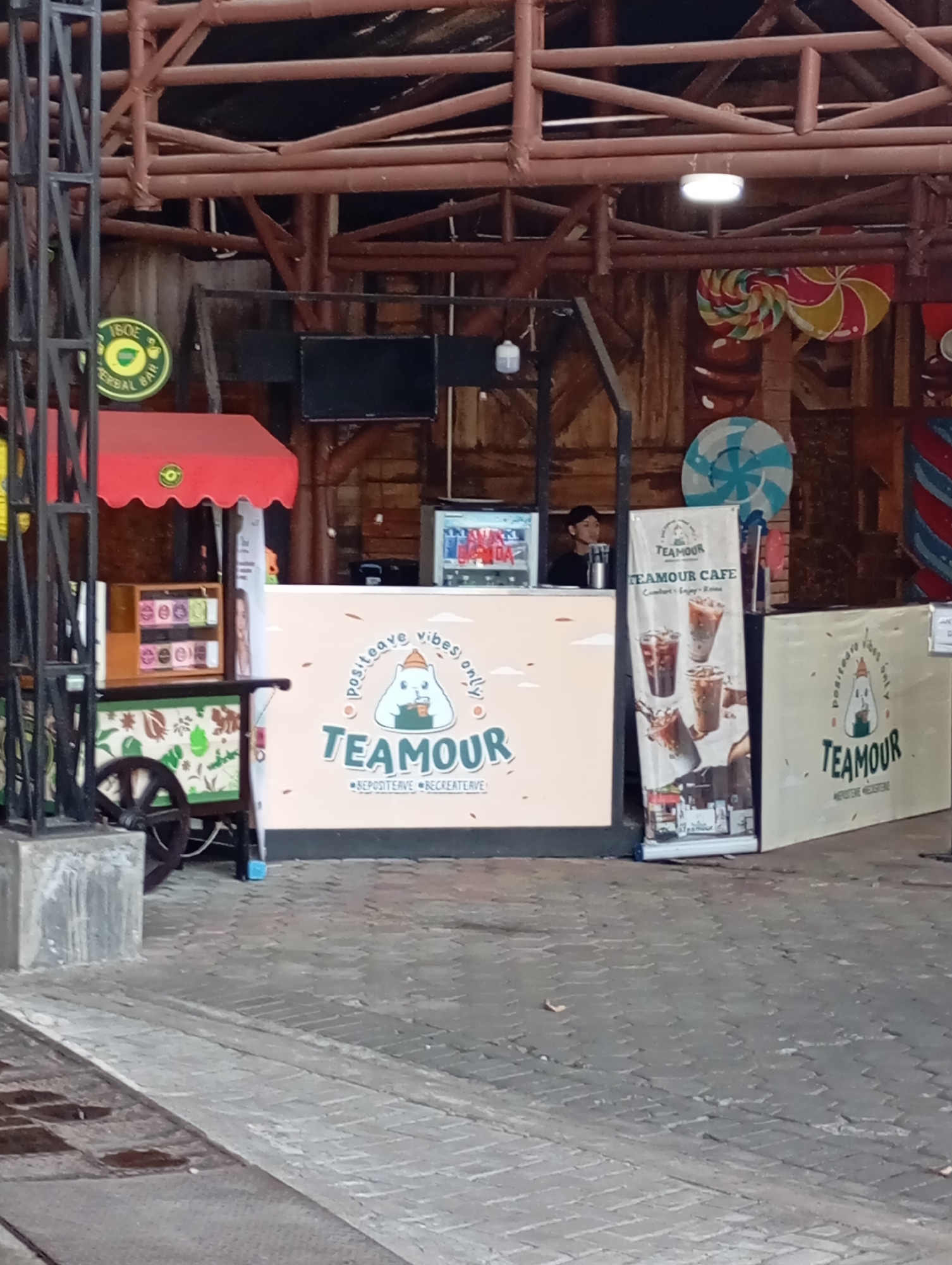 A small, outdoor food stall named "TEAMOUR CAFE" is set up with a light pink and white counter and an adjacent cart filled with colorful packaged goods. The counter features a large banner with images of iced drinks and the brand name prominently displayed. A young person, likely an employee, stands behind the counter. Behind the stall, the scene is decorated with whimsical, oversized candy-like imagery, including a large red and yellow pinwheel and a blue and white pinwheel. The stall is situated on a cobblestone pavement under a rustic, wooden-beam structure, suggesting it might be located in a public market or a themed area within the city of Batu, Indonesia.