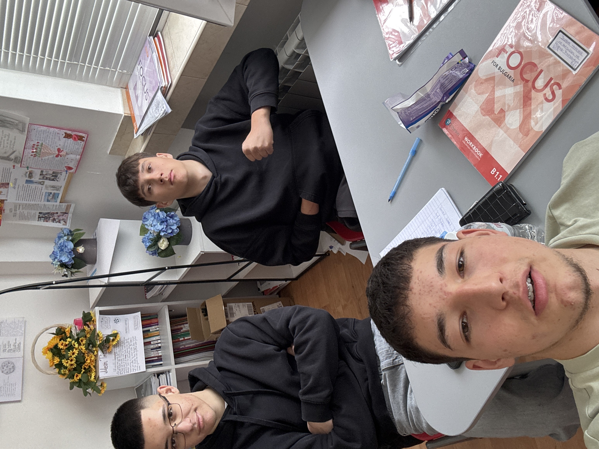 The image captures three young males in what appears to be a classroom or study environment in Zlatograd, Bulgaria. The photo is taken from a perspective looking down at two of the individuals, while the third is in the background, making it seem like a selfie or a casual group photo.
In the foreground, on the right side of the rotated image, is a young male with short dark hair, some facial hair, and braces on his upper teeth, looking directly at the camera with a slight smile. He is wearing a light-colored long-sleeved top. His arm rests on a grey desk. To his left, another young male, also wearing a black hoodie, is visible from the chest up, looking towards the camera. Behind him, partially obscured by a desk, is a third male in a black hoodie, with a short buzz cut and glasses, also looking at the camera with a neutral expression.
The setting suggests a school or study room. Desks are visible, one of which holds a "FOCUS FOR BULGARIA WORKBOOK B1.1" by Pearson, with handwritten Bulgarian text on its cover indicating it's an English workbook for 11th grade, belonging to "Mariya-Magdalena". Nearby are an empty Oreo package and a blue pen. The wall behind the individuals is decorated with various papers, including one titled "Mein Traum" (German for "My Dream") and others mentioning "VETERINARY" and "Architect", possibly indicating future aspirations. A window with white blinds is in the background, letting in natural light, suggesting it is daytime. Shelves holding books, binders, and small boxes are also visible. Decorative elements include artificial blue flowers in pots on a windowsill and yellow artificial sunflowers in a basket on a shelf. The overall atmosphere is relaxed, indicating a break or a casual moment during a study session.