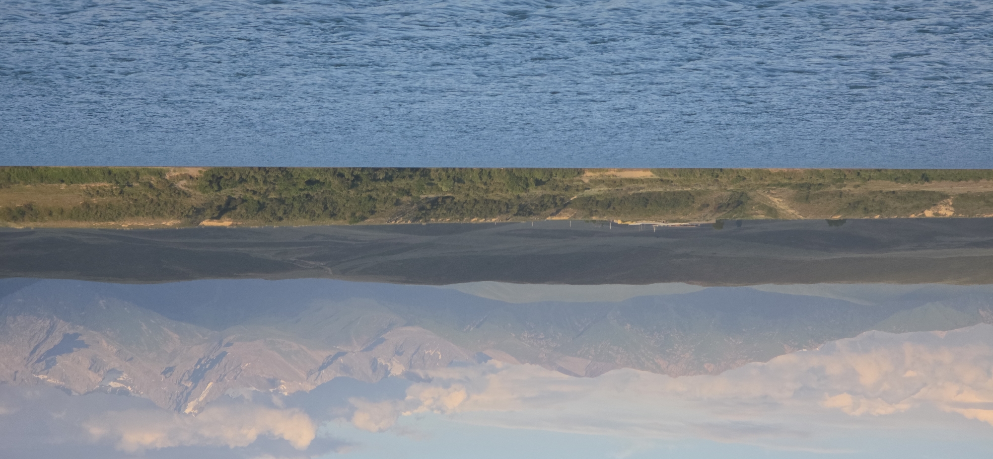 The image captures a serene natural landscape with a body of water in the foreground and mountains in the background, under a soft, cloudy sky. The water is a vibrant blue, rippling gently, suggesting a peaceful atmosphere. Beyond the water, a green, tree-covered slope rises, transitioning into rolling, grassy hills that recede into the distance. These hills are muted in color, indicating a considerable distance. The furthest element of the landscape is a majestic mountain range, its peaks sharp and rugged, with some snow visible on the higher altitudes. The mountains are bathed in the warm, soft light of either sunrise or sunset, casting a gentle glow and creating a hazy, atmospheric effect. A few clouds are present in the sky, tinged with pink and orange from the ambient light. The overall scene evokes a sense of tranquility and vastness, characteristic of remote natural environments. The location is identified as Pristan'-Przheval'sk, Kyrgyzstan, a region known for its stunning natural beauty, including large bodies of water and imposing mountain ranges. No people or man-made structures are prominently visible, emphasizing the natural focus of the image.