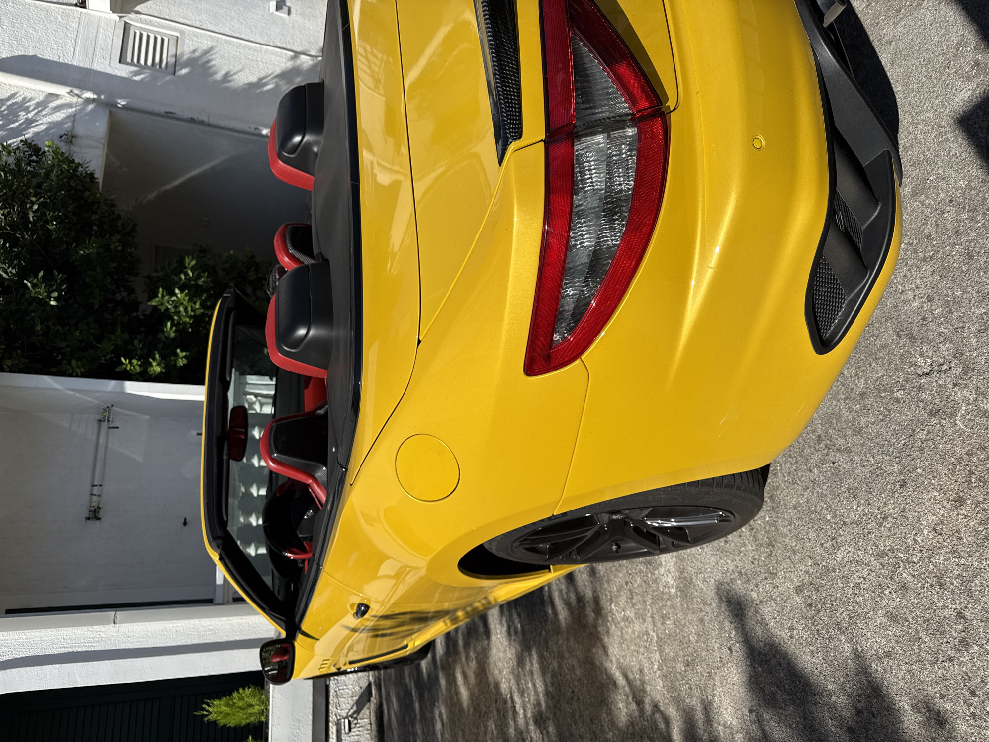 A bright yellow convertible Maserati is parked on a concrete surface in front of a white building with dark green shutters and trees. The car's interior features red leather seats. The rear of the car, including the taillights and a black diffuser, is visible. Shadows from trees are cast across the car and the ground. The location appears to be a driveway or parking area.