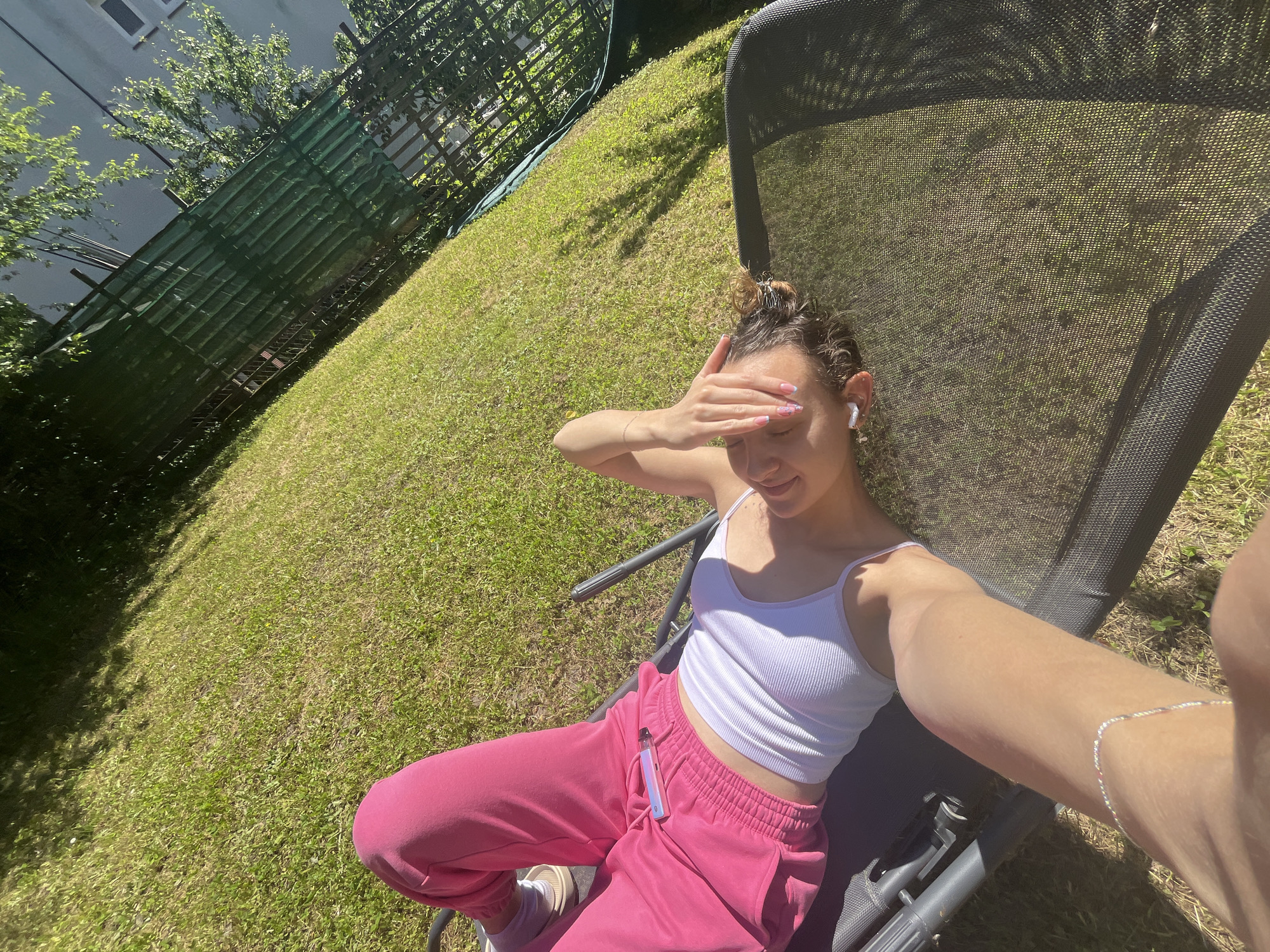 The image shows a young woman reclining on a dark gray mesh lounger outdoors, presumably in Saarbrücken, Germany. She is fair-skinned, with her brown hair gathered in a messy bun, and is wearing a white tank top and bright pink sweatpants. She is facing the camera and appears to be taking a selfie, with her right arm extended. Her left hand is held up to her forehead, seemingly shielding her eyes from the sun. She is wearing an earbud in her left ear and a bracelet on her extended right arm. On her sweatpants, a vape pen is visible. The lounger is placed on a lawn with sunlight dappling through the trees. In the background, there is a green metal fence, a tree, and part of a building.