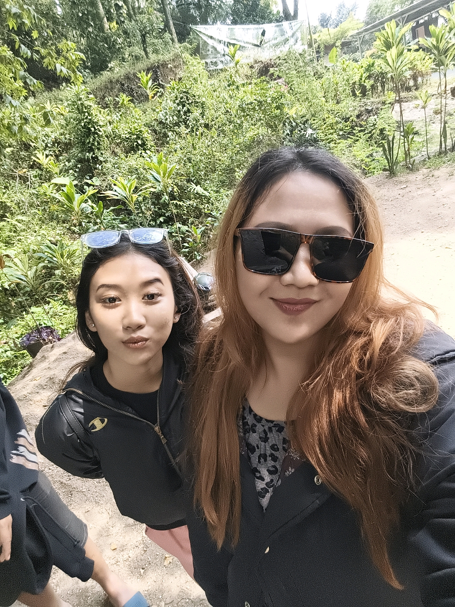 Two young women pose for a selfie outdoors with lush greenery and a dirt path in the background. The woman on the right, closer to the camera, has long, reddish-brown hair and is wearing large, dark-rimmed sunglasses and a dark jacket over a patterned top. The woman on the left, slightly further back, has dark hair with sunglasses perched on top of her head. She is wearing a black jacket. The environment suggests a tropical or sub-tropical location with dense foliage and trees. The path appears to be unpaved, indicating a natural or less developed area. The scene is likely located in Babakan Madang, Indonesia, given the visual characteristics of the landscape.