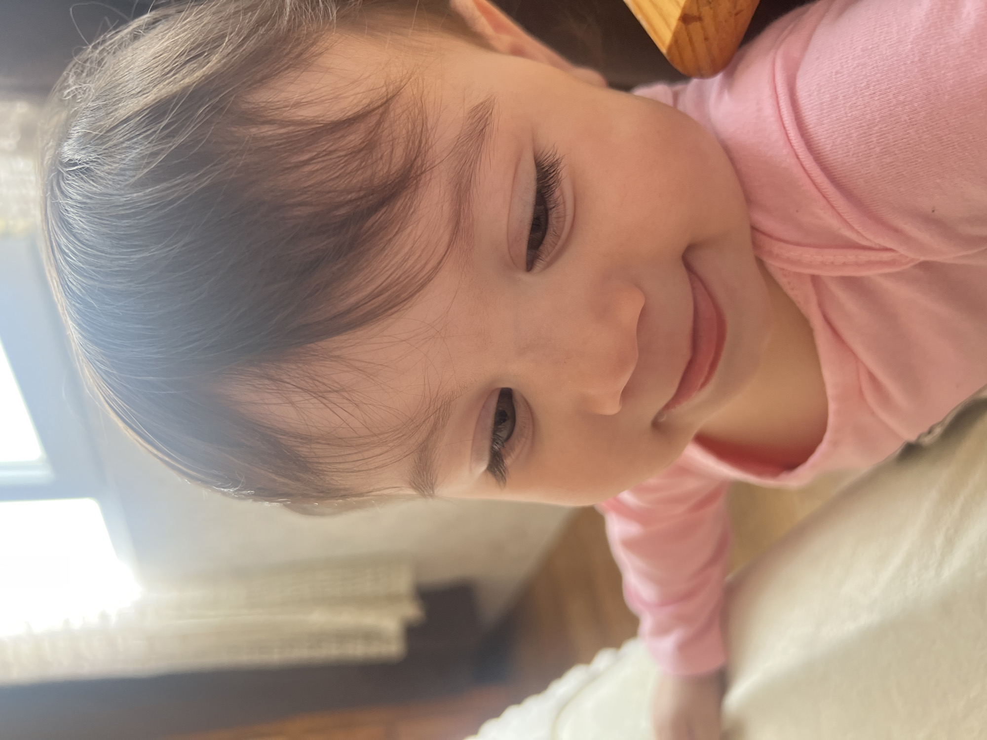 A close-up, slightly tilted, portrait of a baby girl with fair skin, brown hair, and brown eyes. She is wearing a pink long-sleeved onesie. She has a slight smile and her gaze is directed downwards. The background consists of a light-colored, possibly white, surface with some texture, and hints of a window with light filtering through. A wooden element is visible in the upper right corner. The location is unknown and cannot be determined to be Elena, Bulgaria from the visual information provided.