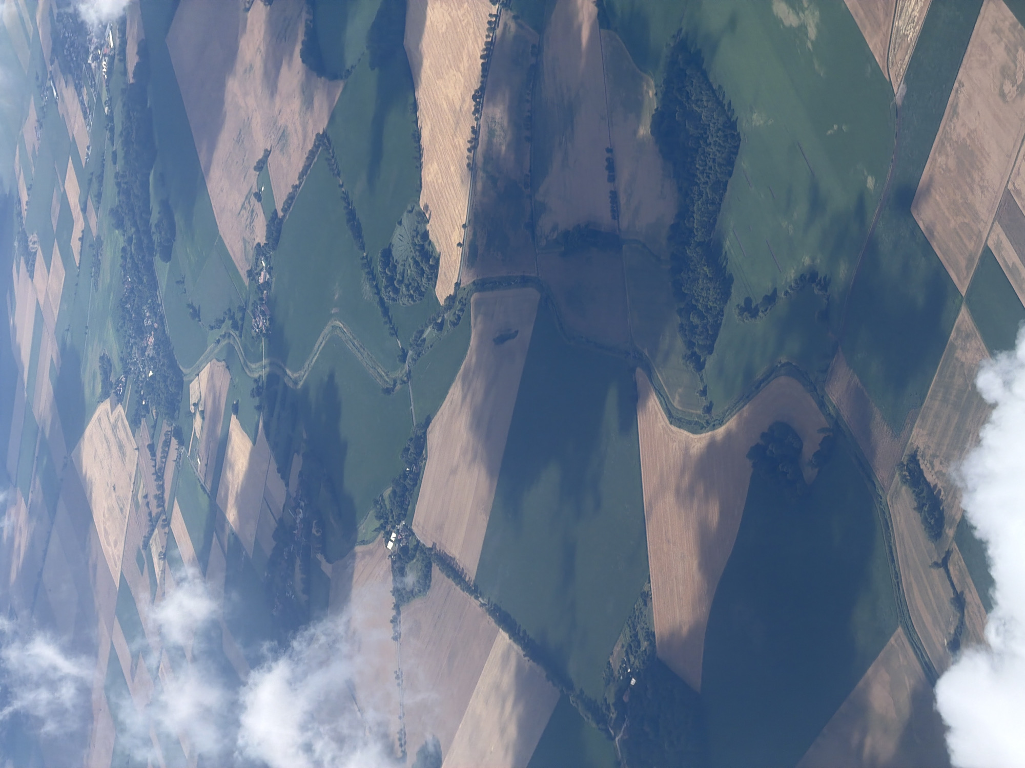 An aerial view captures a patchwork of agricultural fields in various shades of green and brown, dissected by roads and small bodies of water. Clouds drift across the landscape, casting shadows that create patterns of light and dark. The image displays a rural environment with evidence of human activity in the form of cultivated land and scattered buildings. The precise location of Raczkowa, Poland, is not discernable from the visual information presented in the image. There are no people or specific subjects depicted, only the vast expanse of the cultivated land.