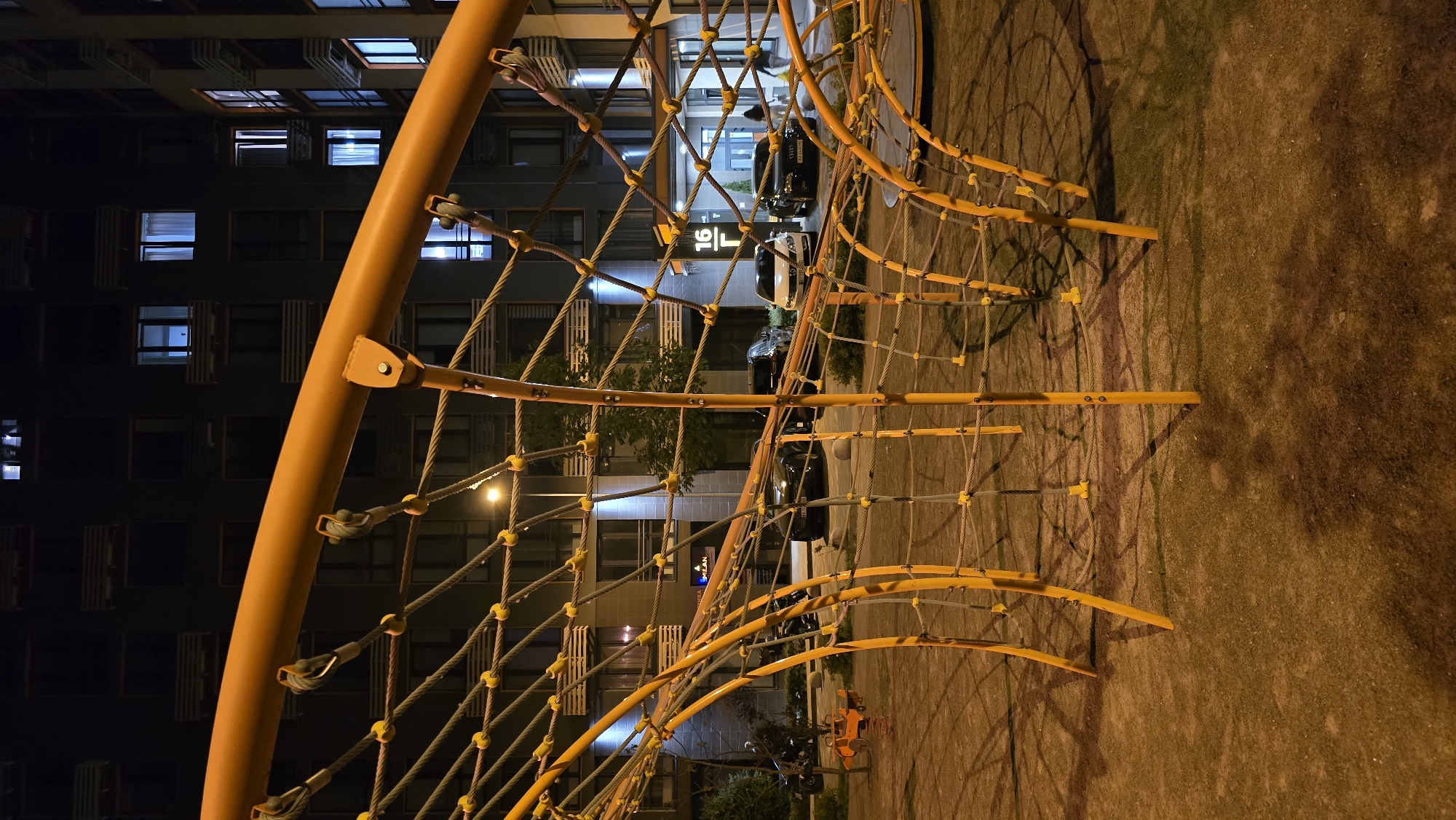 The image presents a nighttime view of a modern playground structure set against an urban backdrop. In the foreground and midground, a prominent, bright orange climbing frame dominates, consisting of thick curved poles and an intricate web of ropes and smaller orange rungs. This structure casts long, complex shadows onto the surrounding grassy ground, which is illuminated by artificial light, appearing in warm tones of brown and muted green.
Behind the playground, a multi-story, dark-colored building rises, featuring numerous windows. Many of these windows are illuminated with a cool, bluish light, indicating occupied residential or office spaces. Several cars are parked along a street or driveway in front of the building. Scattered trees and foliage are also visible, hinting at some landscaping.
The time of day is clearly night, characterized by the dark sky and the reliance on artificial lighting from the building windows and streetlights. No specific weather conditions are discernible. The setting appears to be a residential area or a public park within an urban environment, consistent with Kyiv, Ukraine.
A notable detail is a sign on the building displaying the clear text "16 L". Above this, another sign with indistinct white text on a dark background is partially visible. No people or active events are taking place, resulting in a quiet and still atmosphere. The striking orange of the playground equipment provides a strong focal point against the darker hues of the night sky and the building.