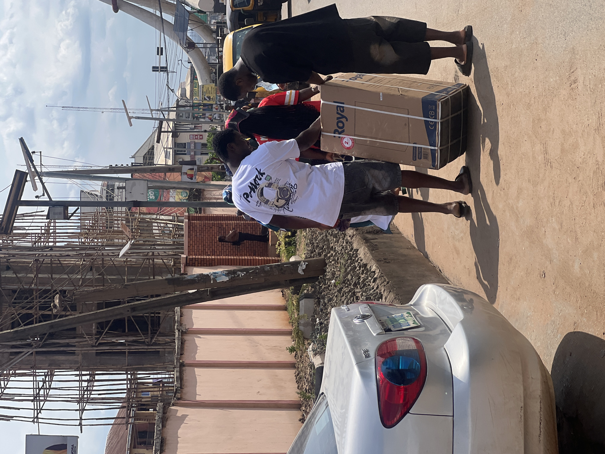 The image depicts an active street scene in an urban area of Onitsha, Nigeria, under bright daylight. Three men are the primary subjects, focused on transporting a large, rectangular cardboard box along a paved surface, likely a sidewalk. The box appears heavy and is being carried by two men in the foreground, with another man in a dark shirt assisting from behind. The man on the left in the foreground wears a white t-shirt with a graphic and the word "Pollock" printed on it, paired with dark shorts and sandals. The second man wears a red and black striped shirt. Their shadows are cast prominently on the ground, indicating the sun is high.
In the foreground, the rear of a silver car is partially visible, parked on the side of the road. Its blue-tinted taillights are a noticeable detail, and part of its license plate reads "IMO SO" and "NIGERIA". The background is bustling with elements typical of an urban environment: numerous utility poles with crisscrossing wires, a tall communications mast, and multi-story buildings. One building on the left appears to be under construction or renovation, covered in wooden scaffolding. Other buildings display various commercial signs, including one with "EASY WAY" visible.
The cardboard box being carried has several visible texts, including "Royal", "2", "105 LITERS", and symbols like "CE CB". The sky above is partly cloudy. The overall atmosphere suggests a normal daily activity in a vibrant city environment.