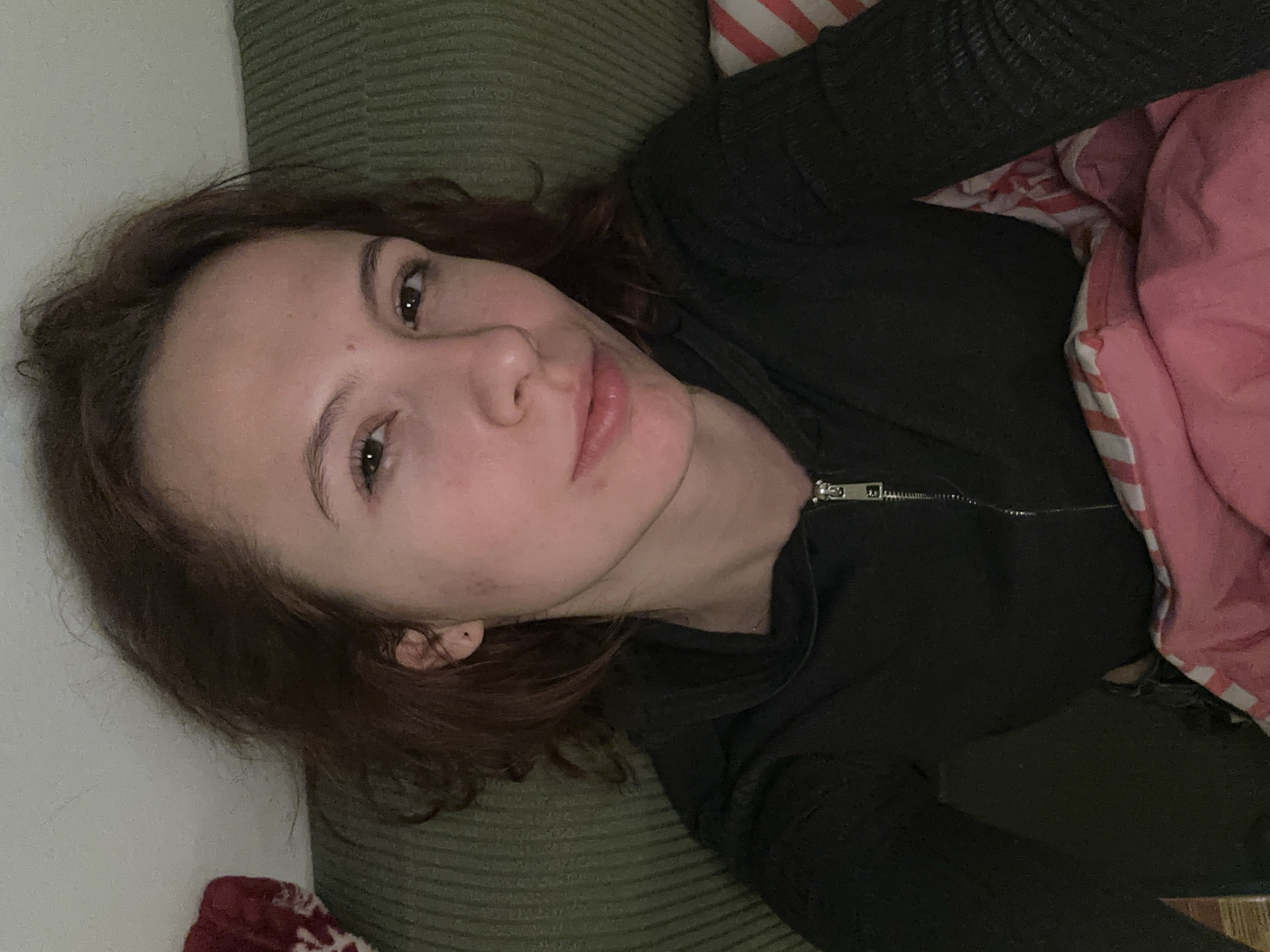 A woman with brown hair and fair skin is lying down. She is wearing a black zip-up top. She is partly covered by a pink and white striped blanket. She is lying on a green textured surface, possibly a couch or bed. A white wall is visible. The location is not discernible from this image, and there is no indication it is in Novi Sad, Serbia.