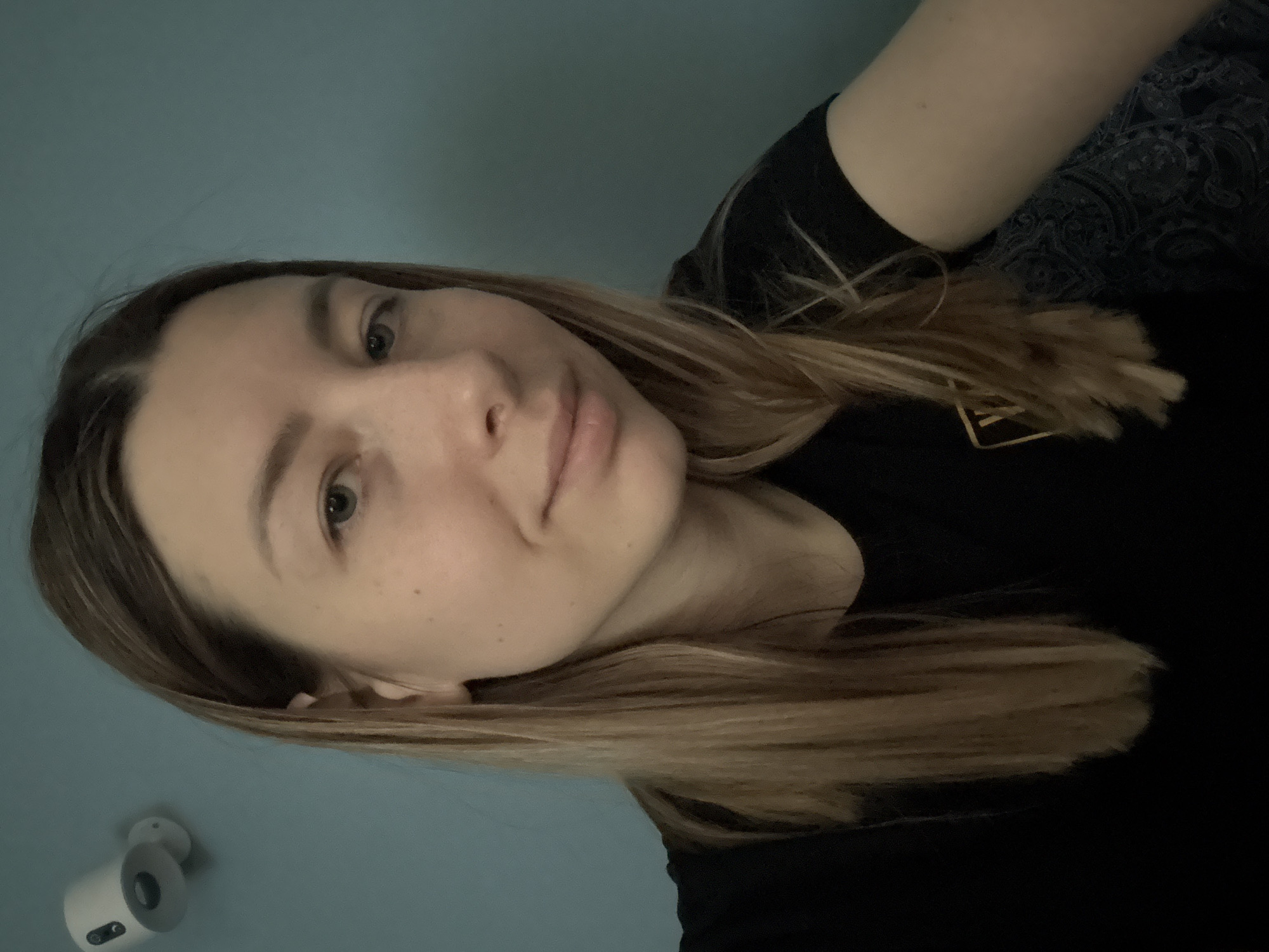 A woman is pictured indoors, facing the camera. Her hair is a blend of light brown and blonde. She is wearing a black shirt. A security camera is visible in the lower left corner of the frame. The wall behind her is a light blue color. A patterned fabric is seen in the background, near her right shoulder. The city of Moscow, Russia cannot be confirmed as the location.