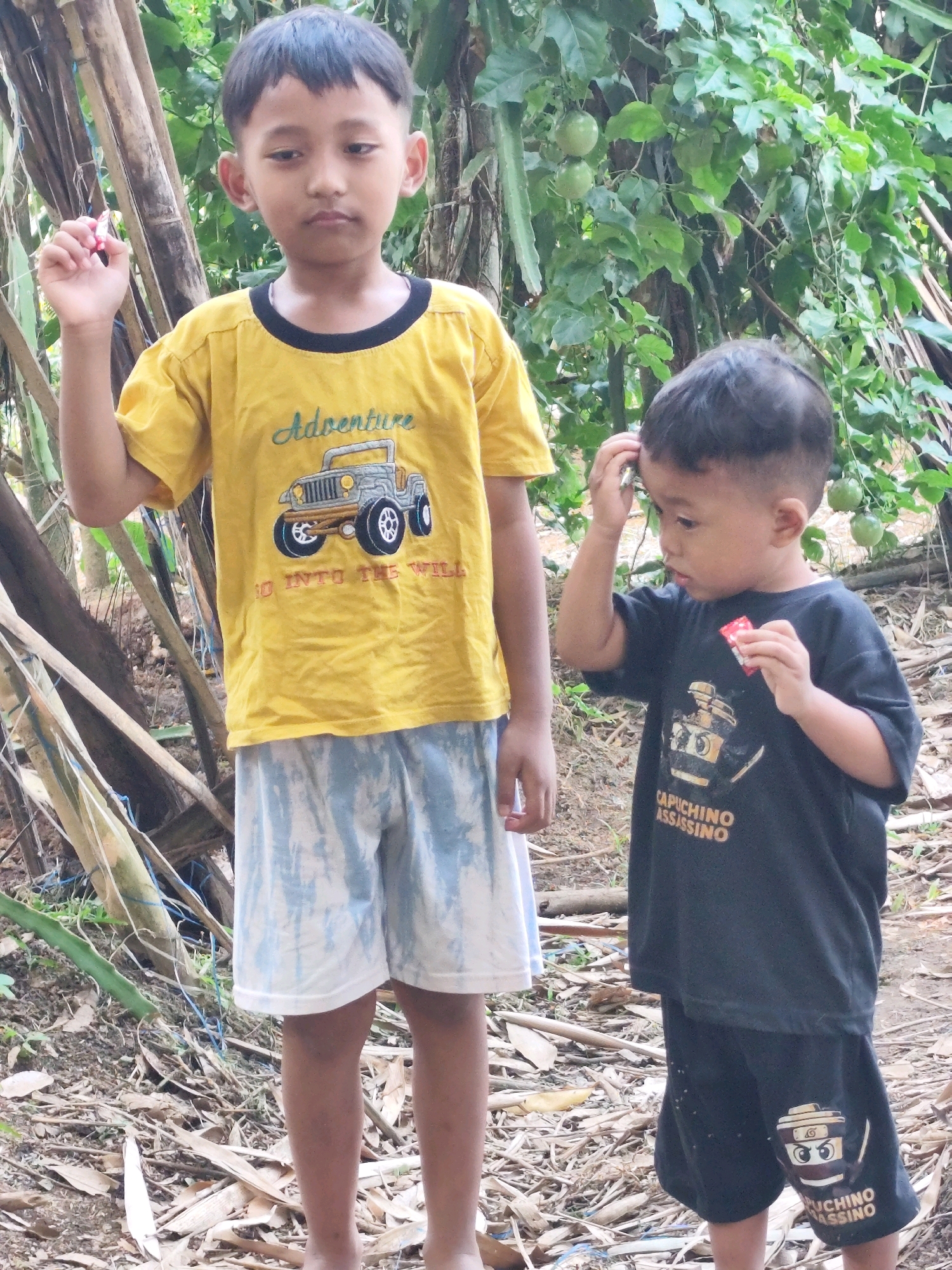 Two young boys stand in a yard with dry leaves and branches scattered on the ground. The boy on the left, wearing a yellow t-shirt with a graphic of a jeep and the word "Adventure" and "Go Into The Wild," holds a small red and white object in his left hand. He has dark hair and is looking down. The boy on the right, wearing a black t-shirt and black shorts with graphics and text including "CAPUCHINO ASSASSINO," has his right hand on his head and is holding a small red object in his left hand. He is looking to his left. Behind them, a vine with green fruit hangs from a tree. The scene appears to be outdoors in a rural or semi-rural setting, possibly in Jambewangi, Indonesia, given the vegetation and the overall environment.