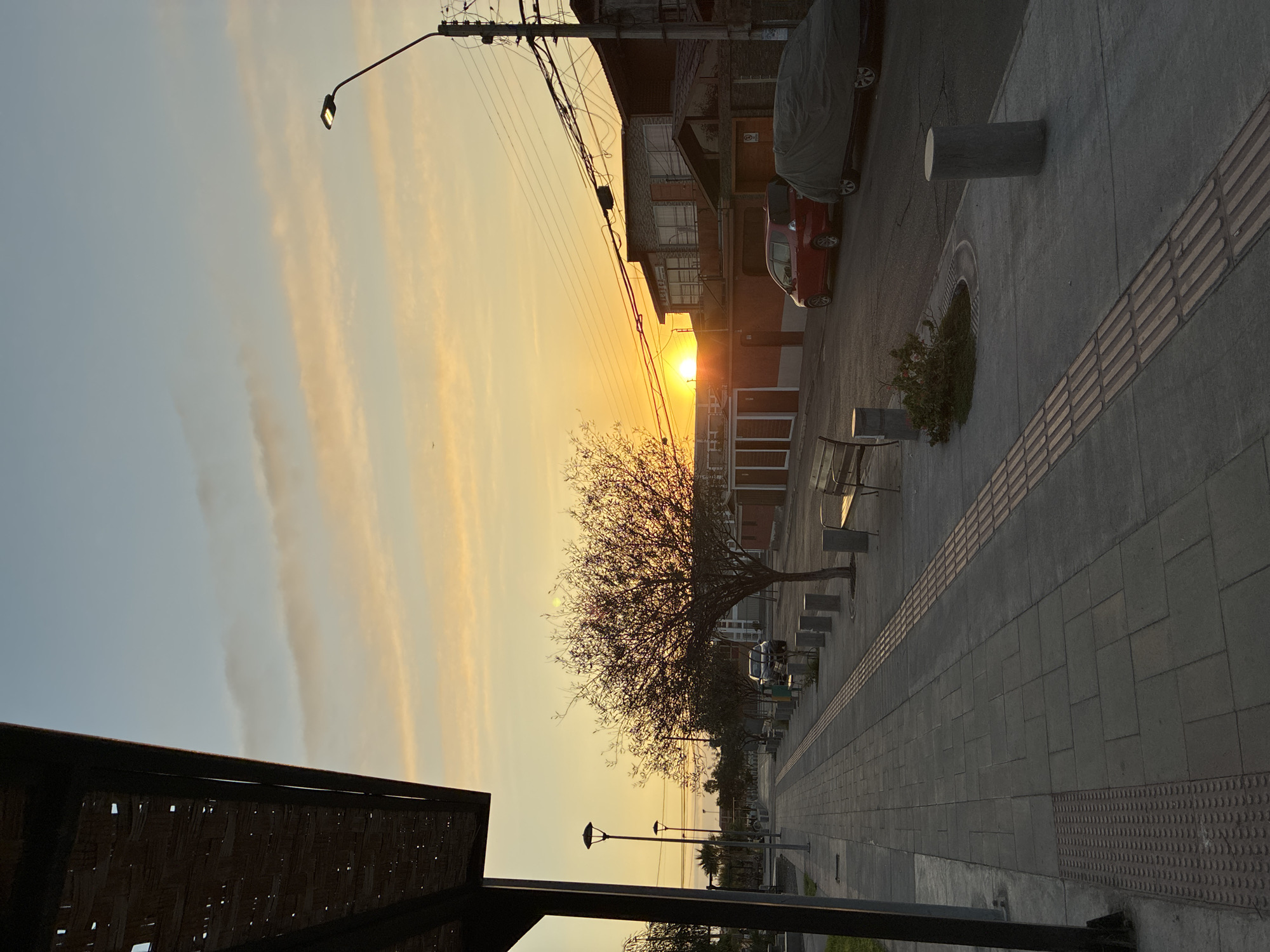 The image shows a street scene in Arica, Chile, during sunset. The sun is setting behind a building, casting a golden light across the sky and street. A bare tree stands prominently in the foreground, silhouetted against the bright light. The street is paved with grey tiles and features a tactile paving strip for visually impaired pedestrians. Several benches line the sidewalk, along with a few cylindrical bollards. A red car is parked along the street, partially covered by a car cover. Overhead power lines and streetlights are visible. The overall environment suggests a quiet, urban setting during the evening.