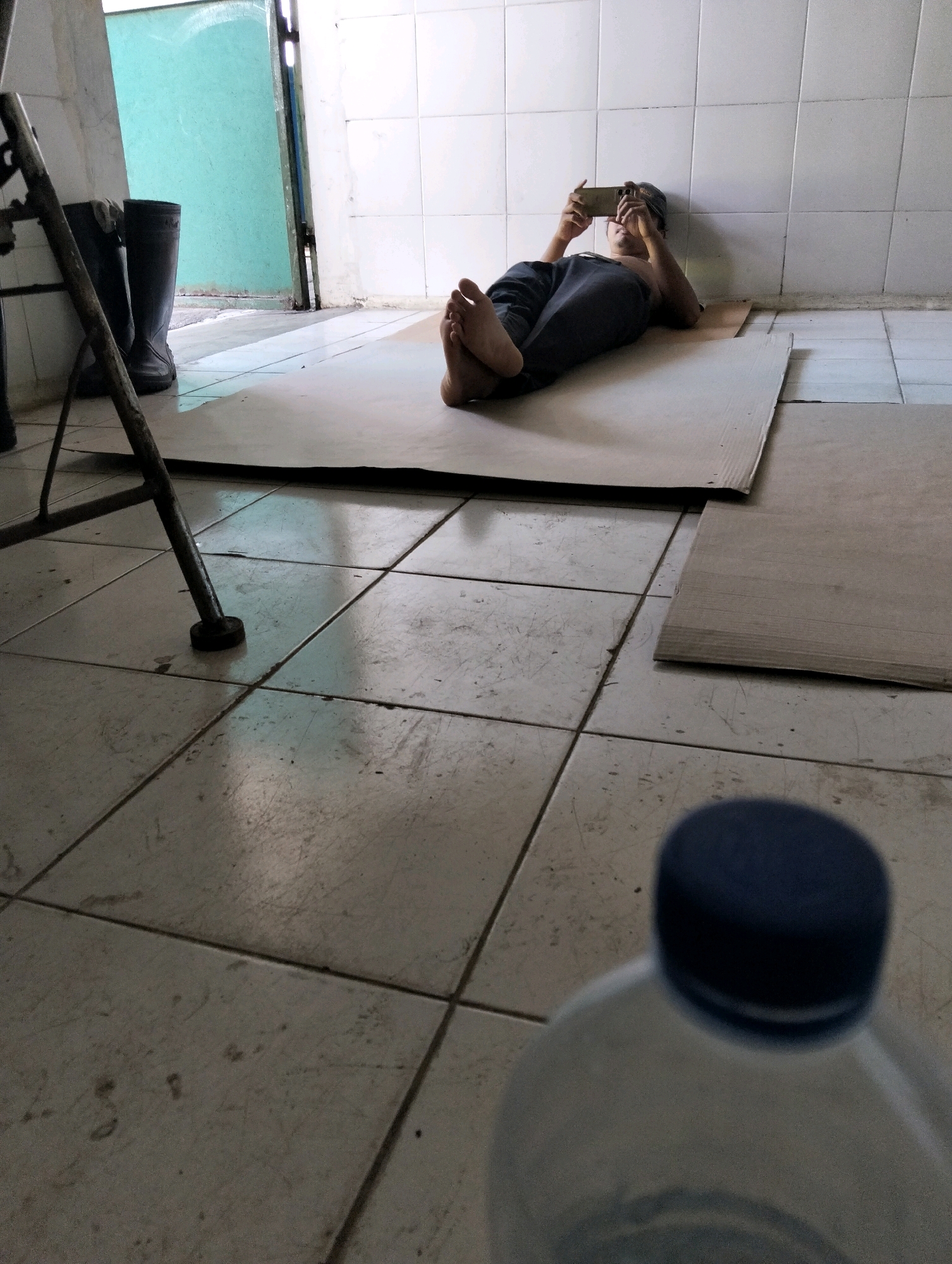 The image depicts an indoor scene, likely within a building in Tanggulangin, Indonesia. The floor is tiled with light-colored square tiles. In the foreground, the blurred top of a clear plastic bottle with a dark blue cap is visible.
The middle ground shows a person lying on their back on a piece of cardboard placed on the floor. They are wearing dark pants and a dark cap. The person is holding a mobile phone horizontally in their hands, looking at the screen. Their feet are visible, extended towards the left of the frame.
To the left of the person, leaning against a wall, are two black rubber boots. A metal stand or frame, possibly part of a piece of furniture or equipment, is also visible in this area, with a metal leg resting on the tiled floor.
The background shows a wall tiled with white square tiles. To the far left, a doorway or opening reveals a green-colored wall. The lighting appears to be coming from an overhead source, casting some shadows on the floor. The overall impression is one of a simple, perhaps utilitarian, indoor space.