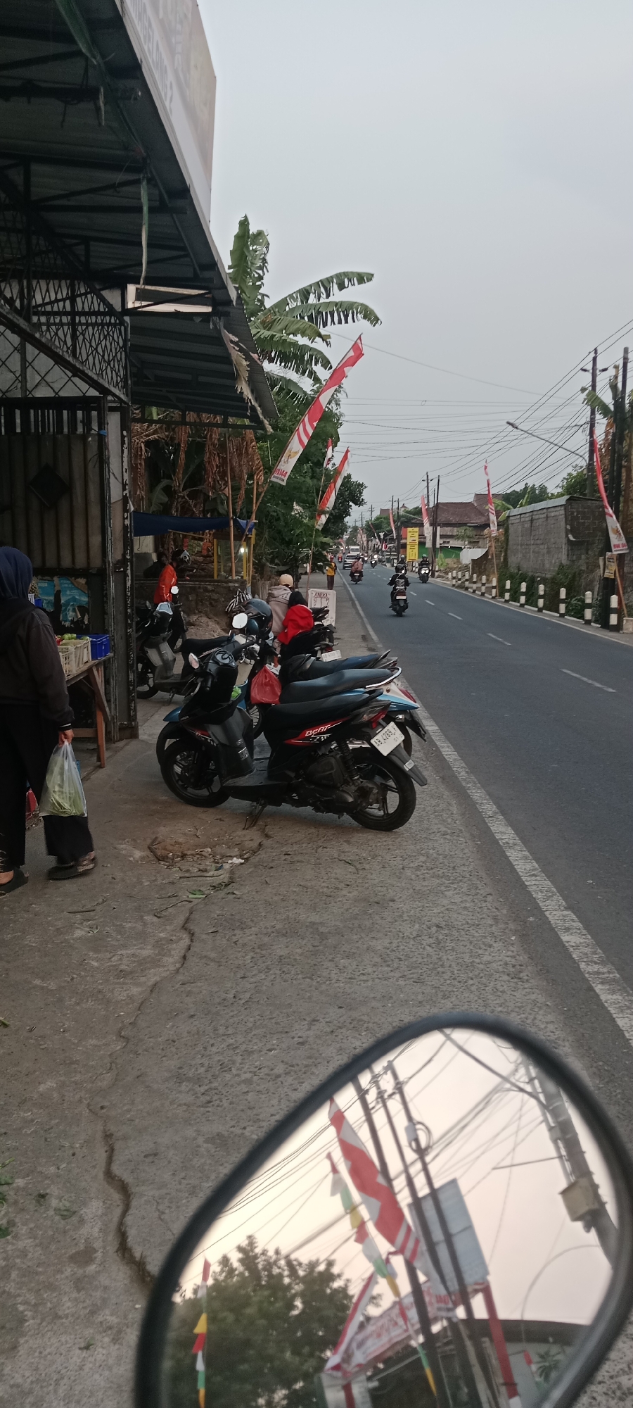 This is a low-angle, medium shot taken from a motorcycle's side mirror, capturing a street scene in Maguwoharjo, Indonesia. The perspective is slightly tilted, with the mirror framing the lower portion of the image. The mirror reflects red and white Indonesian flags, trees, power lines, and a building.
The primary scene, viewed directly, shows a street with a sidewalk. On the left, a person wearing a dark hijab and black pants is standing, holding a clear plastic bag filled with green produce. Adjacent to them is a table with more produce displayed in a blue crate. Behind this is a structure with a metal roof supported by a metal frame, and what appears to be a fence or gate.
Several motorcycles are parked on the sidewalk, facing towards the street. One person in an orange shirt is seated on one of the motorcycles, while another person in a red jacket is also near the motorcycles.
The street itself is asphalt and appears to be a busy thoroughfare, with multiple motorcycles and at least one car visible in the distance. Red and white Indonesian flags are strung along poles on both sides of the street, indicating a celebration or national holiday. The sky is overcast, suggesting early morning or late afternoon. The environment appears to be a roadside market or commercial area. The ground on the sidewalk is concrete, showing some cracks and wear.