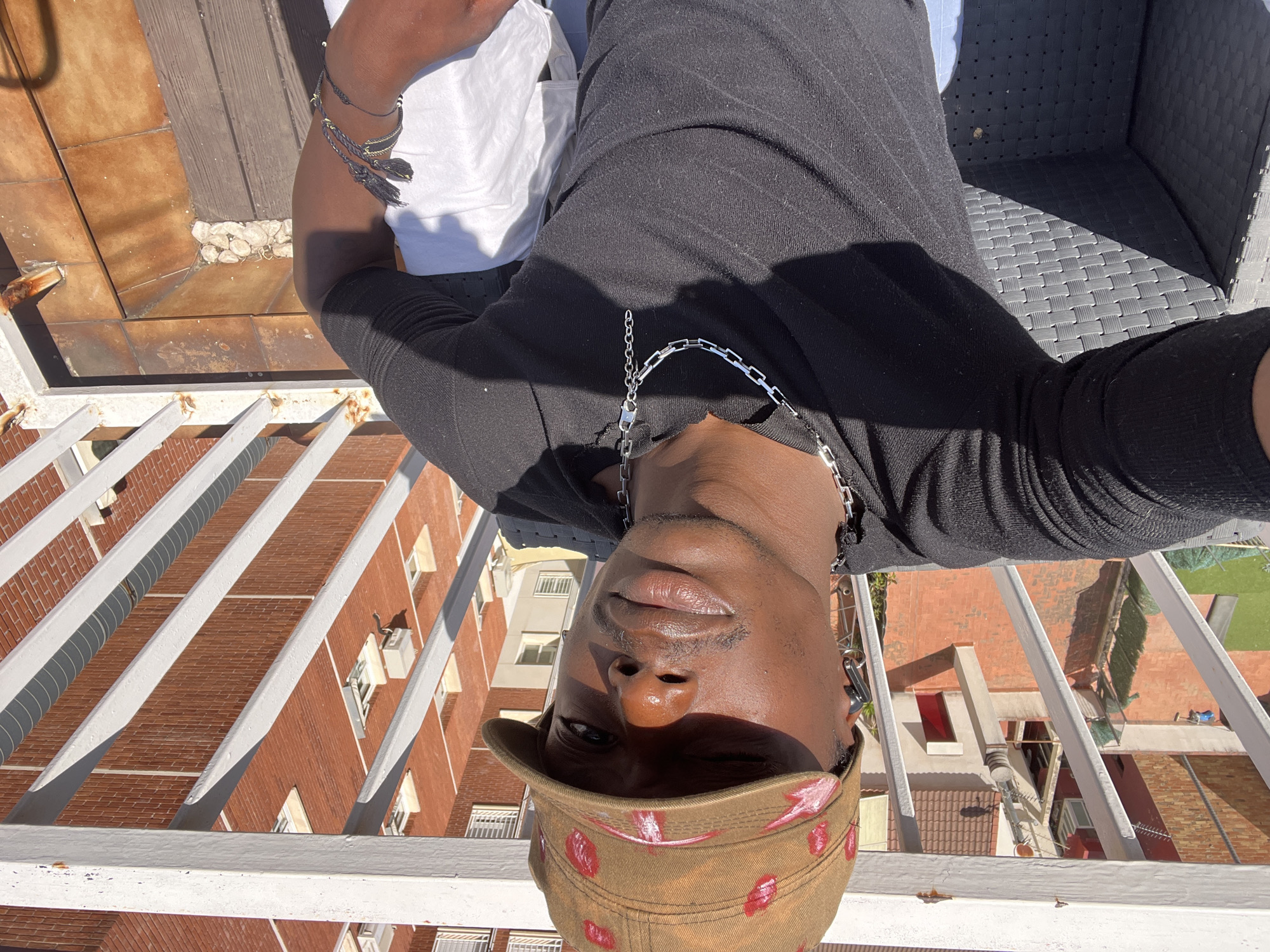 The image is a vertically oriented selfie taken from a high vantage point, looking down onto an urban scene. The subject, an individual with dark skin, is centered in the frame. They are wearing a black long-sleeved top, a silver chain necklace, and multiple braided bracelets with silver and gold accents on their left arm. A distinctive brown cap, adorned with pink and red lip-print patterns, is worn on their head, casting a shadow over their eyes. Their expression appears neutral.
The person seems to be resting or seated on a balcony or rooftop terrace. A white metal railing runs across the foreground and along the left edge of the image. To the subject's right, parts of a black woven outdoor chair and a white pillow are visible.
Below the vantage point, an urban landscape unfolds under bright, clear sunlight. The background features several multi-story buildings constructed with red brick facades and white window frames. Many buildings exhibit traditional orange-red tiled roofs, characteristic of the region. A tiled patio area, likely terracotta, with small white stones, is directly below the railing. Glimpses of green vegetation are visible further in the distance.
The strong sunlight and sharp shadows indicate a sunny day, likely during the morning or late afternoon. The architectural style, particularly the red brick and tiled roofs, is consistent with the provided location context of Barcelona, Spain, portraying a Mediterranean urban environment. No discernible text is present in the image.