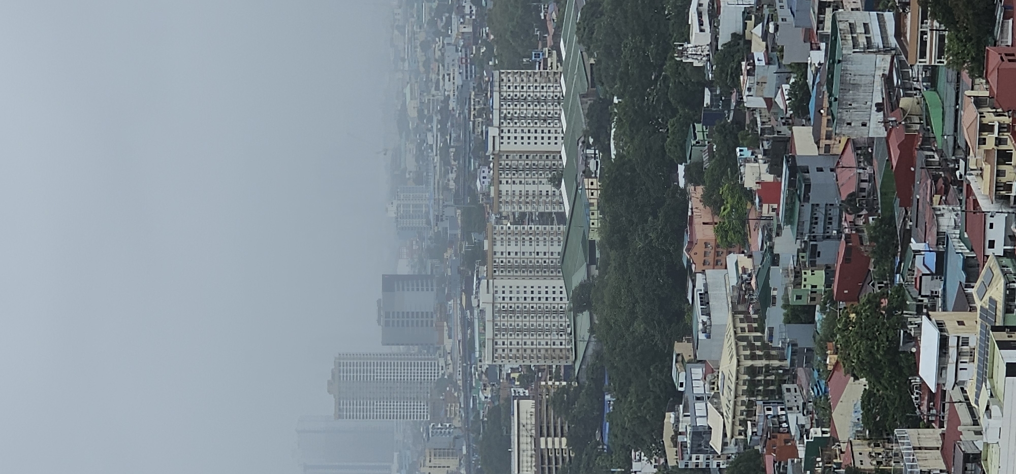This is a vertical, full-frame, eye-level, outdoor, daytime shot of the city of Makati, Philippines, on a hazy day. The camera is looking over a densely populated urban area characterized by a mix of residential and commercial buildings of varying heights and architectural styles. In the foreground and mid-ground, there are numerous low-rise buildings with flat roofs, some featuring visible solar panels and air conditioning units. Many of these structures are painted in a variety of colors, including white, beige, orange, red, and green. Trees are interspersed throughout the urban landscape, particularly in the mid-ground, providing a stark contrast to the built environment.
Further in the mid-ground and background, taller buildings emerge, including several distinct high-rise residential towers. One prominent building in the mid-ground is a pale beige, multi-story structure with a grid of windows. To its left, a taller, light-colored apartment building features a repeating pattern of balconies and windows. In the background, more high-rise buildings are visible, but they are partially obscured by the haze, giving the impression of a sprawling cityscape. The overall atmosphere is subdued due to the overcast or hazy conditions, with muted colors and a lack of sharp detail in the distance. No people are visible in the frame, and the scene depicts a static urban environment.